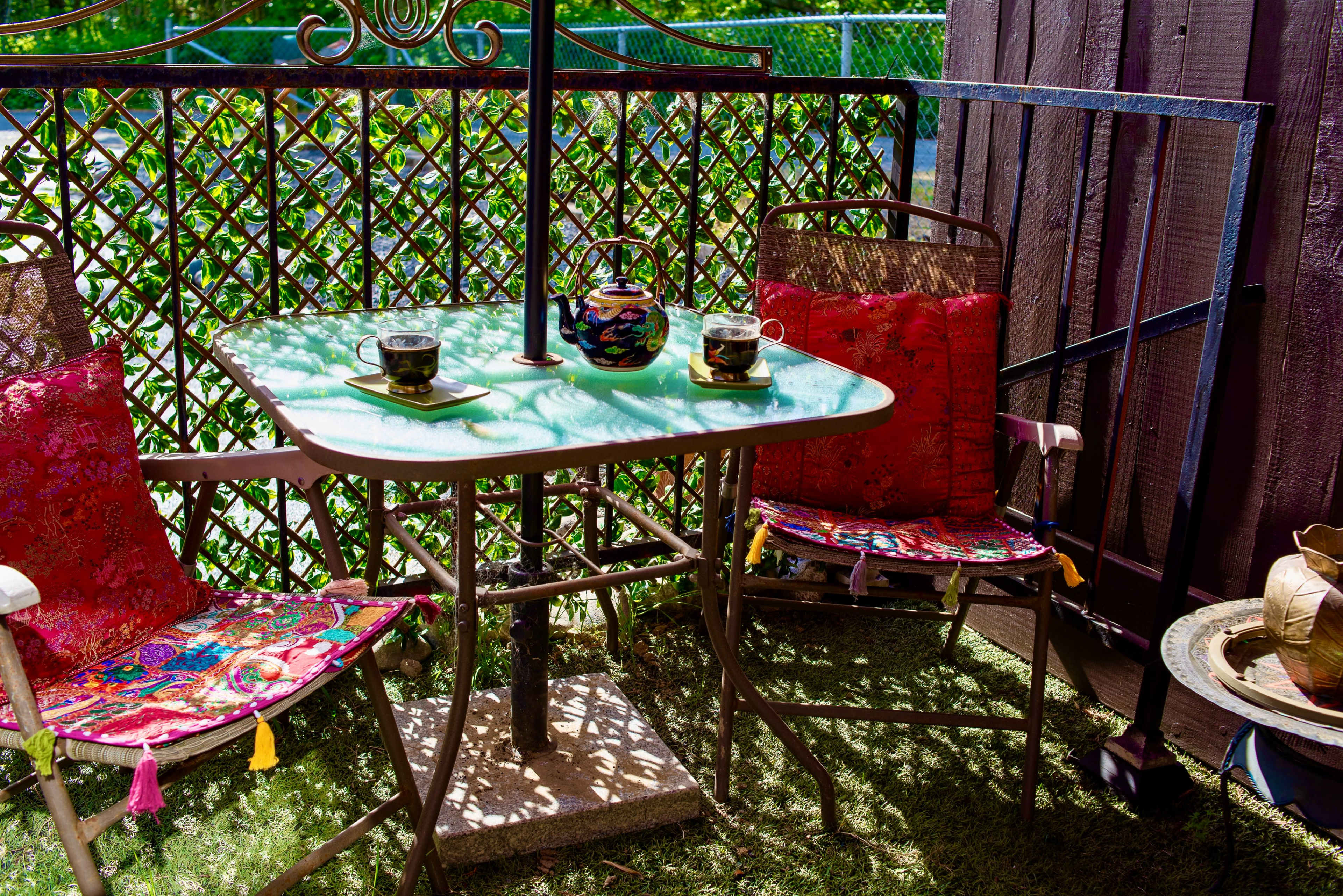A small outdoor table with two metal chairs, each adorned with colorful cushions, is set with a teapot and two cups on a sunlit balcony surrounded by a lattice fence.