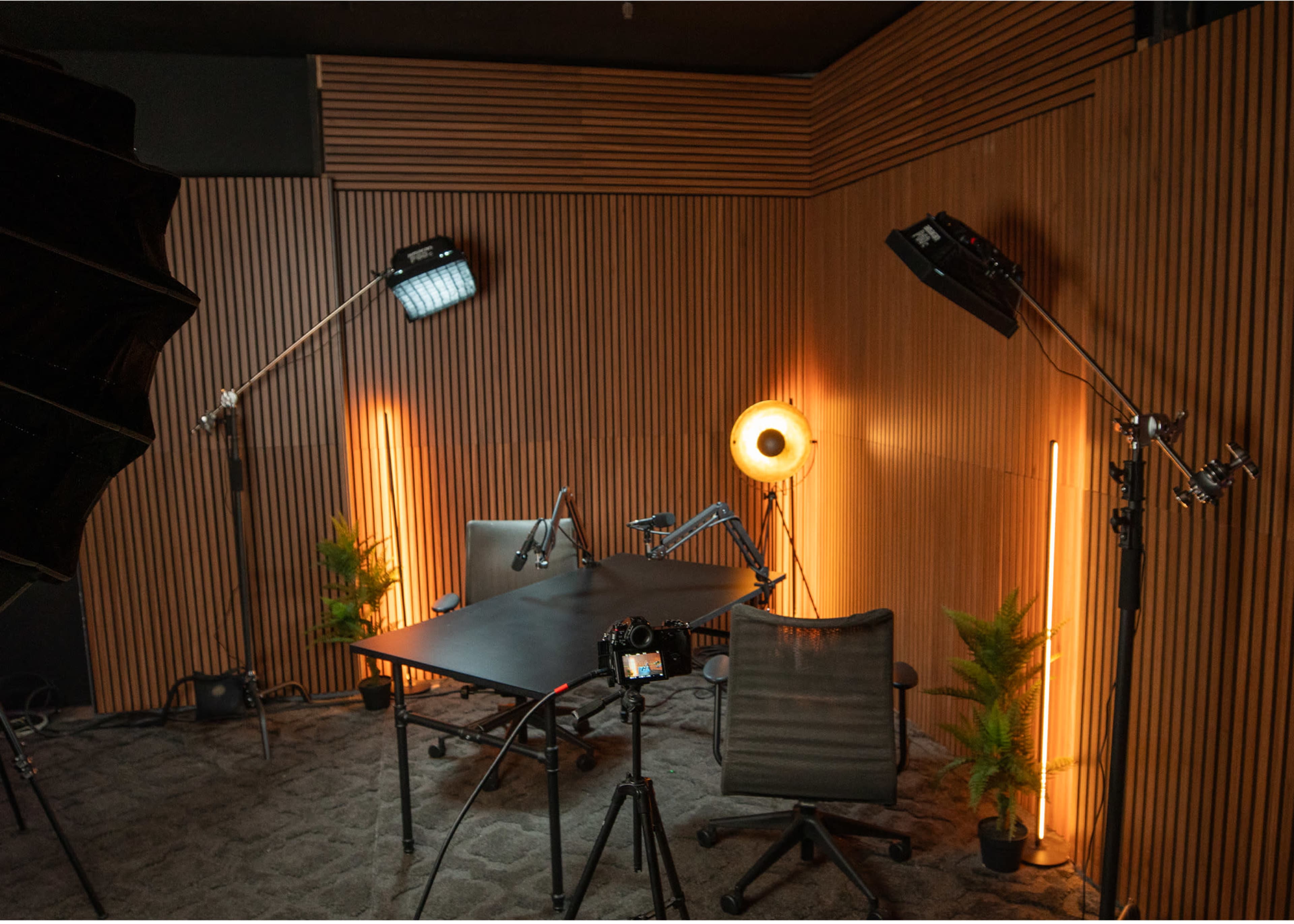 The image shows a well-equipped recording studio featuring a black table, two chairs, studio lights, and a wooden accent wall.