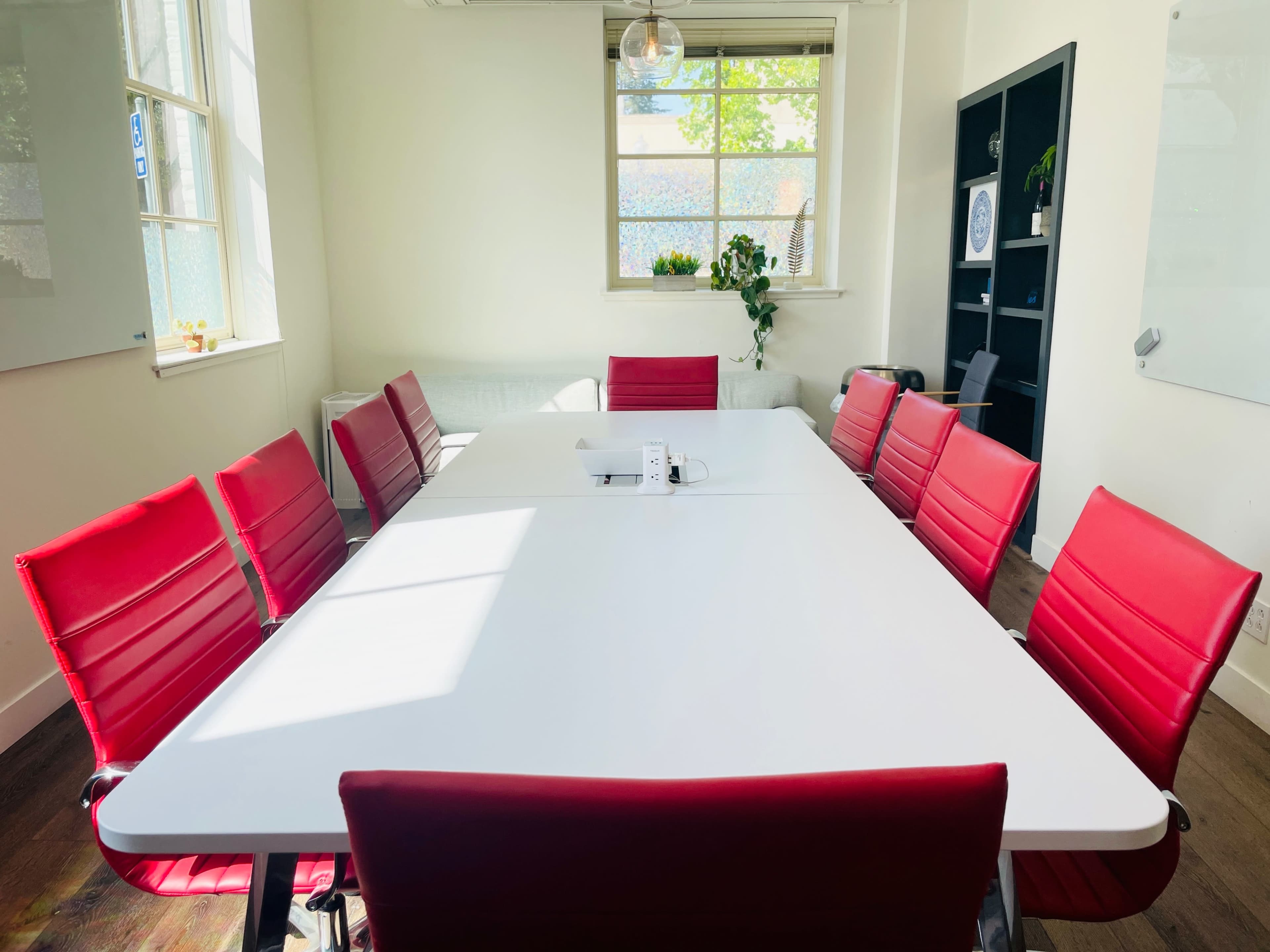 A bright meeting room features a large white table surrounded by red chairs, with sunlight streaming through a window.