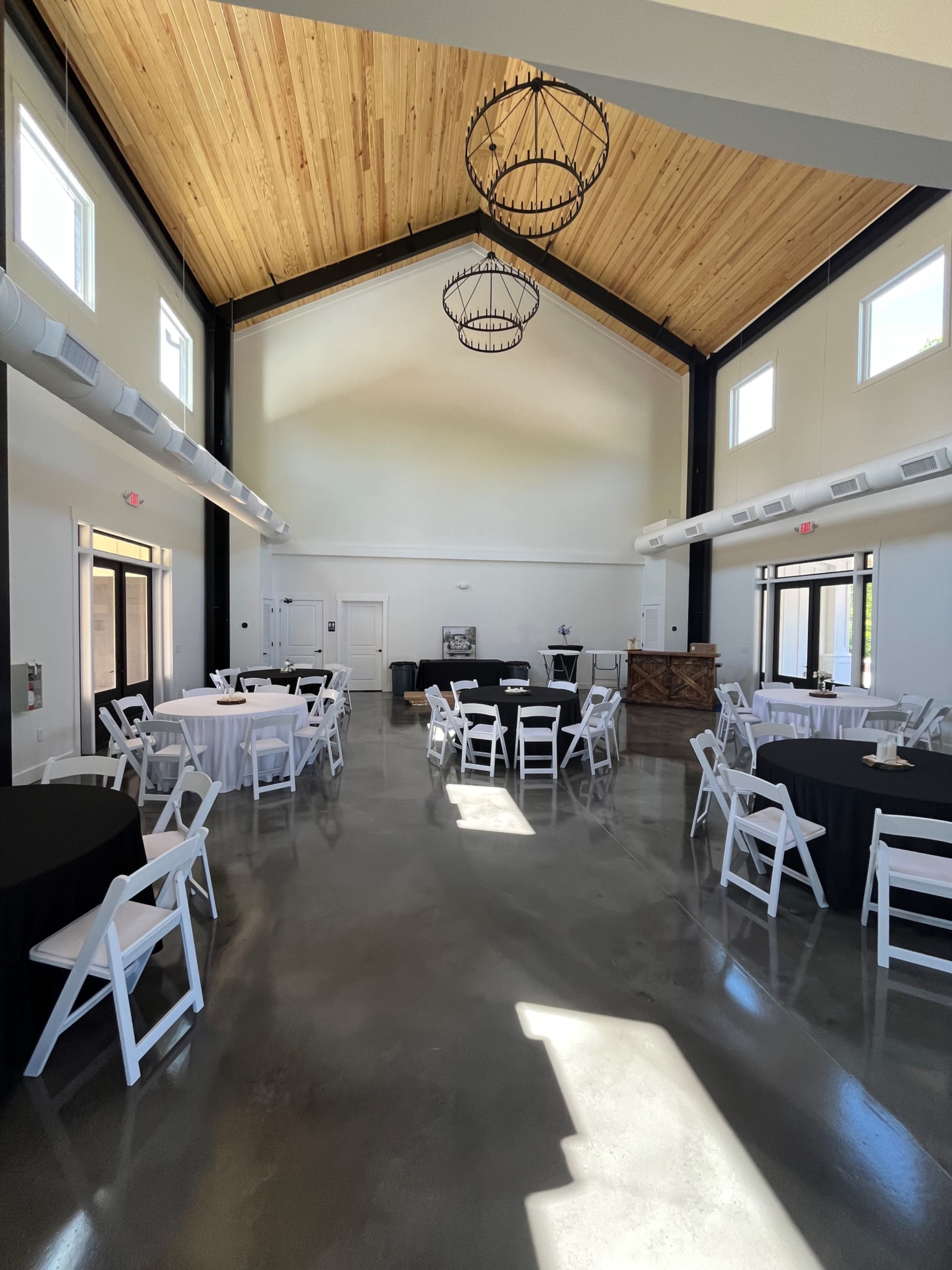 An indoor event space with round tables covered in black and white tablecloths, set beneath a high wooden ceiling with industrial-style light fixtures.