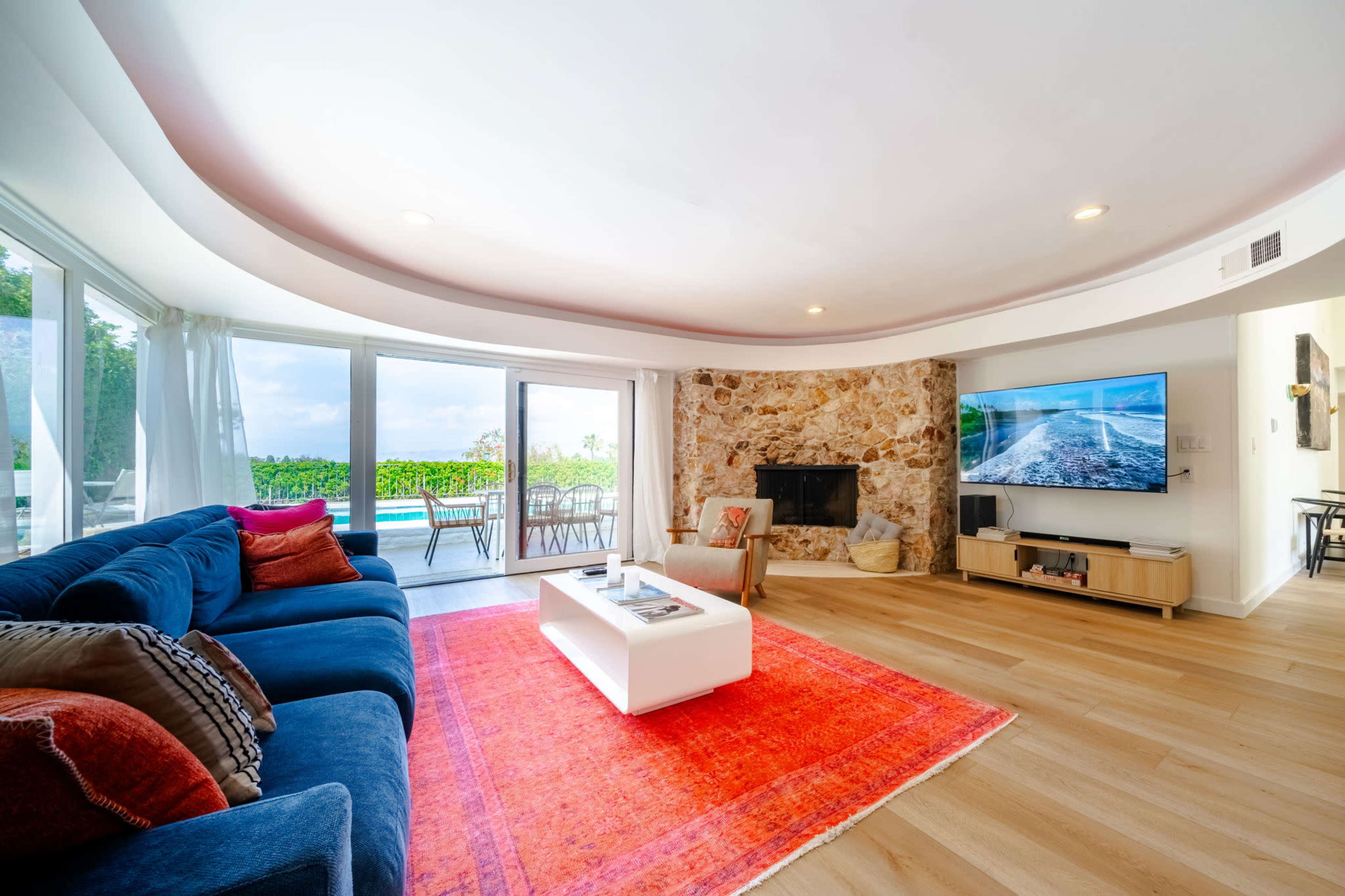 A modern living room features a blue sectional sofa, a white coffee table, and a stone fireplace, with large windows overlooking a pool area.