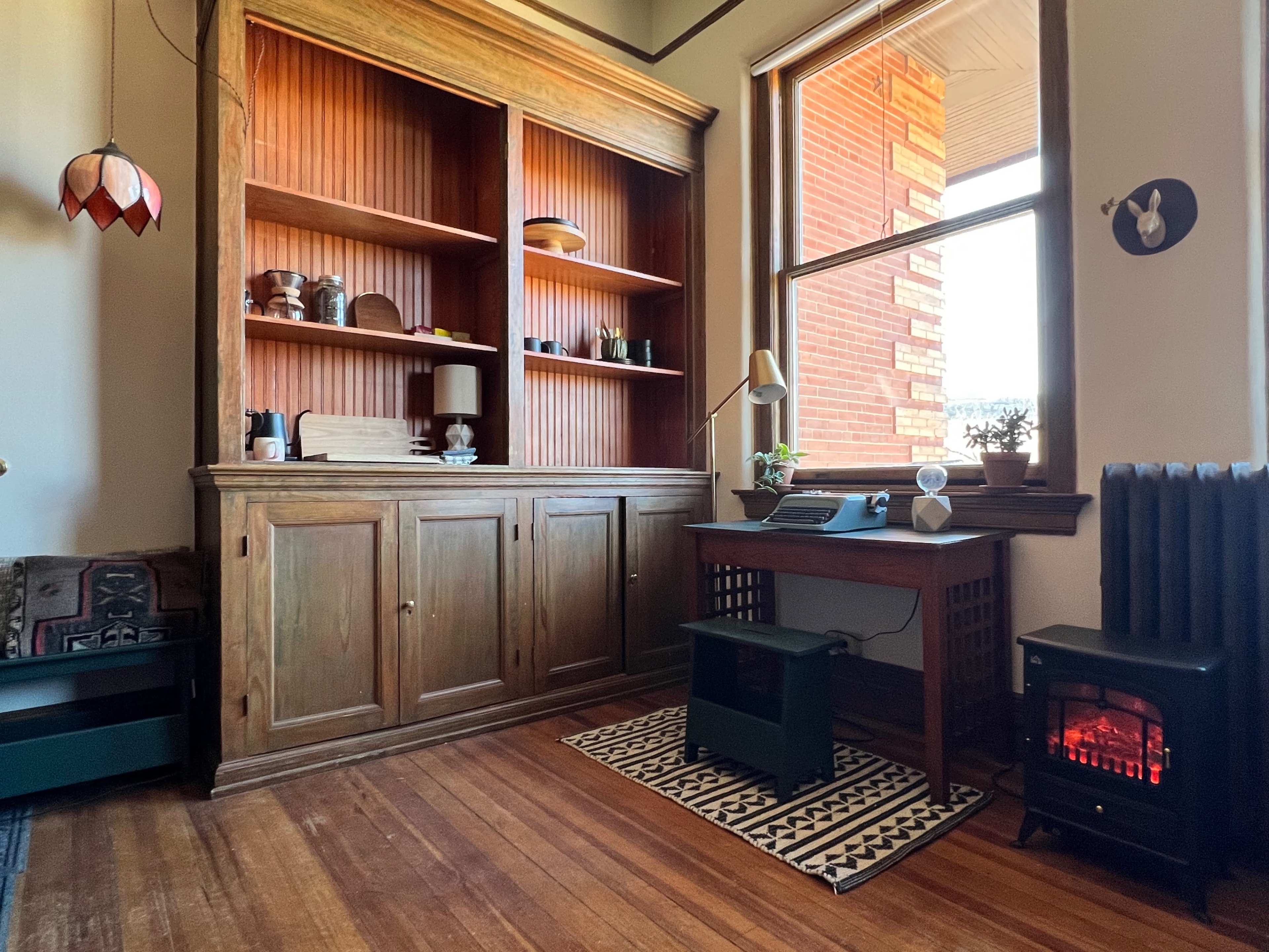 A cozy room featuring a wooden cabinet with shelves, a small desk with a lamp, and a decorative electric heater next to a window.