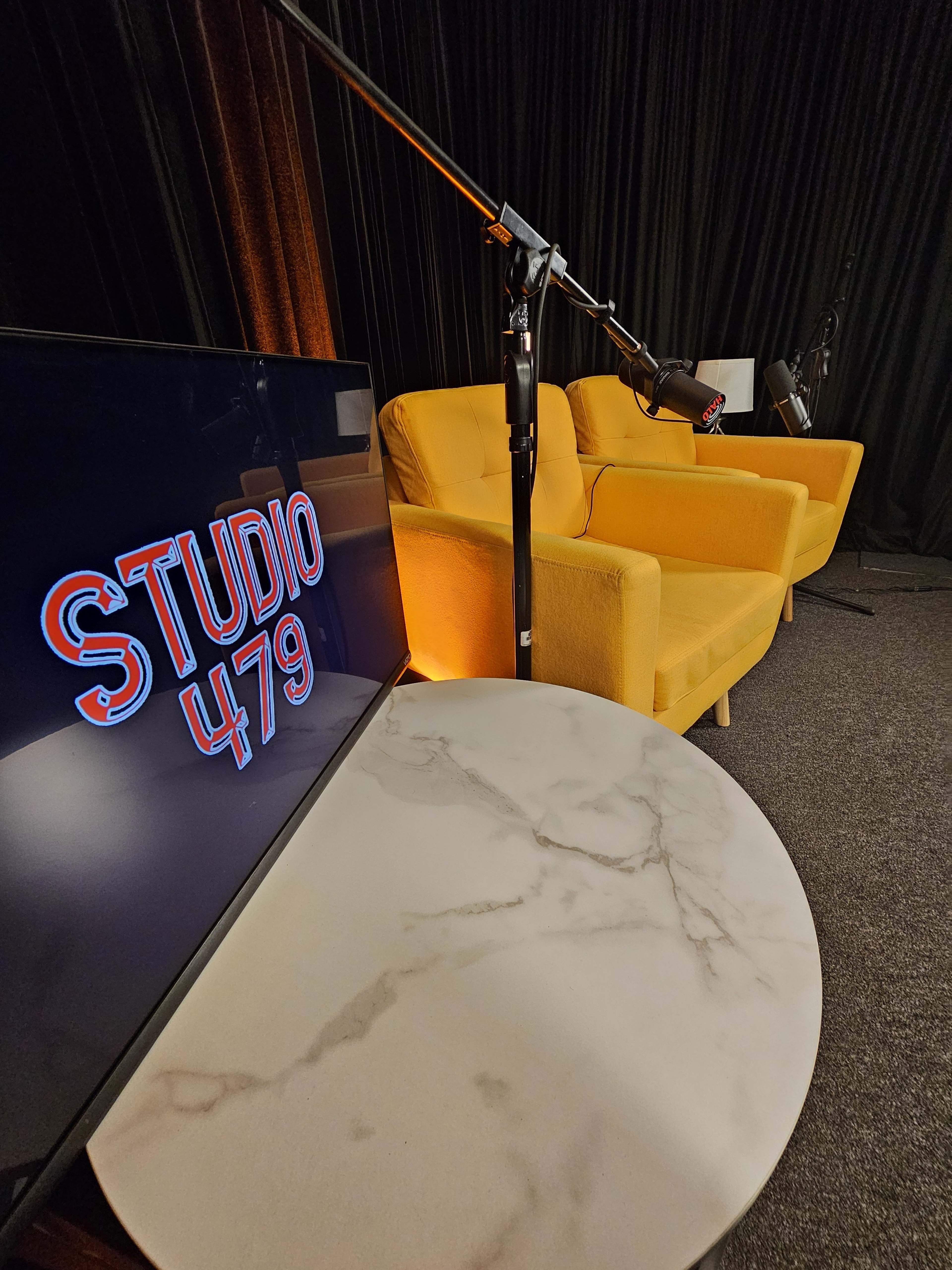 A television screen displaying "STUDIO 479" sits in front of two yellow armchairs, with microphones set on stands nearby, against a dark backdrop.
