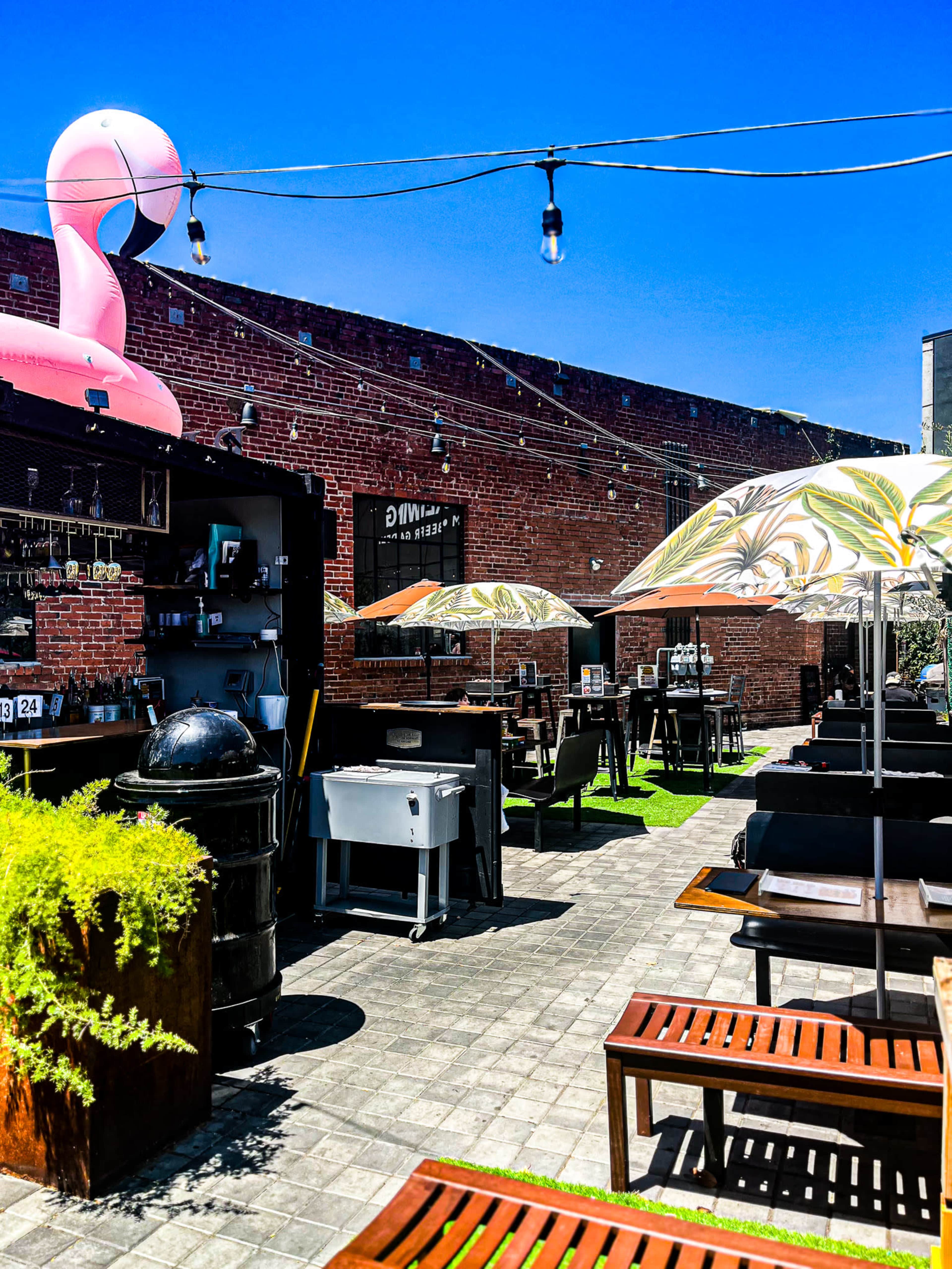 The scene features a vibrant outdoor dining area with decorative umbrellas, grill stations, and a large pink flamingo inflatable against a red brick wall under a clear blue sky.