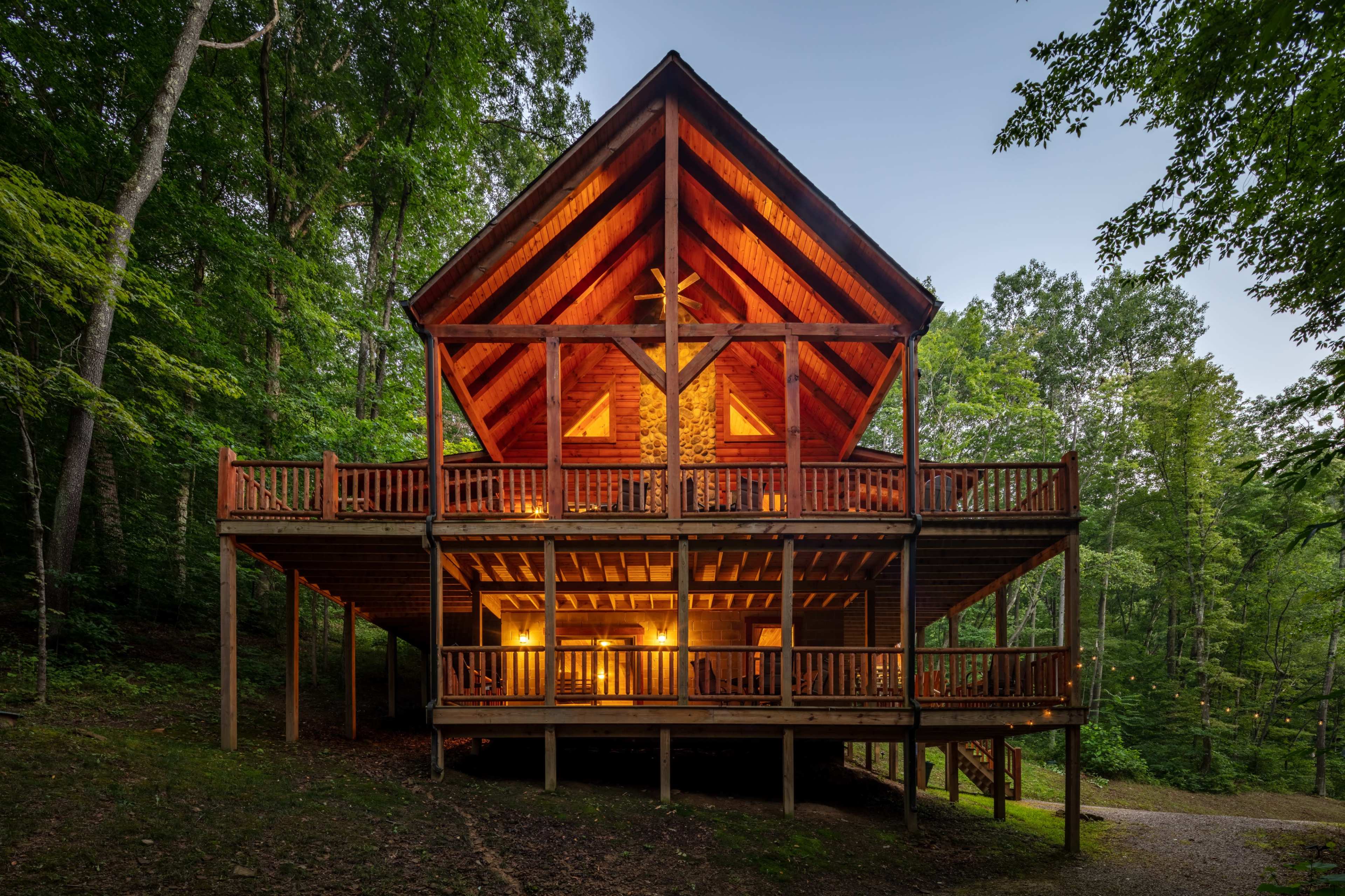 A large wooden cabin with a steeply pitched roof is situated in a forested area, featuring multiple balconies and warm lighting visible from the lower level.