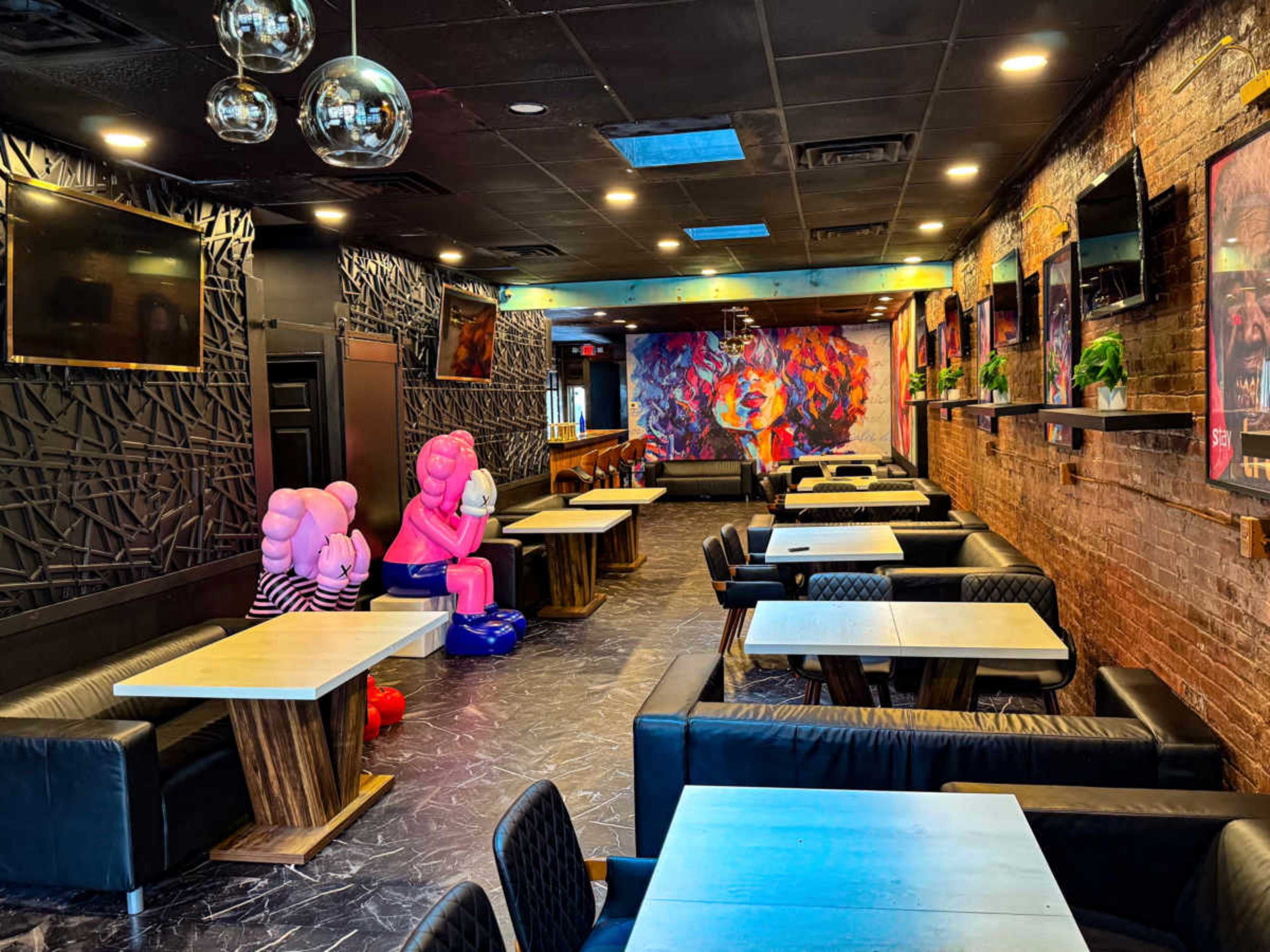 The image shows a stylish restaurant with dark walls, modern furniture, and colorful artwork adorning one wall, along with large seating areas and playful decorations.