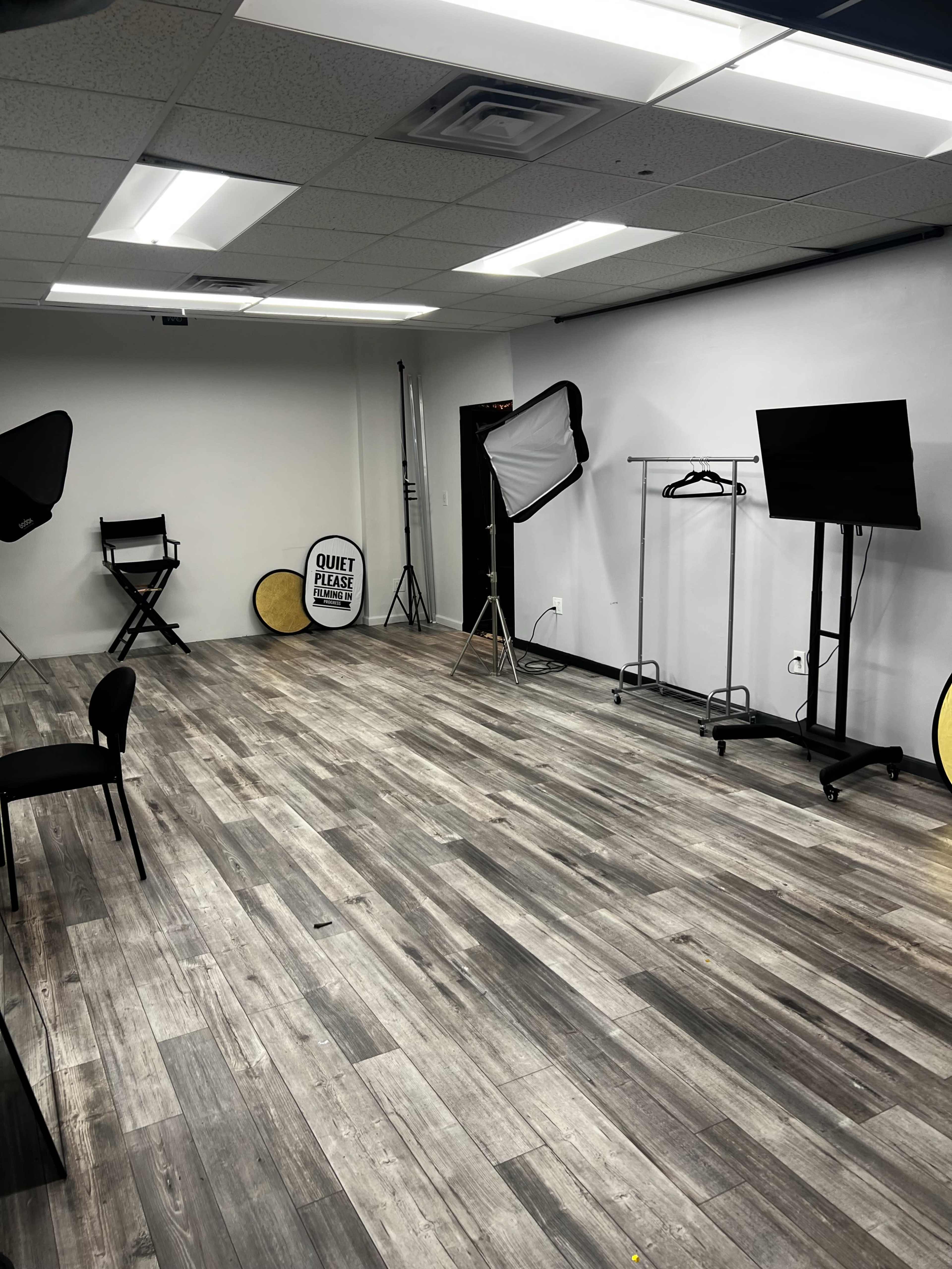 The image shows an empty studio space with wooden flooring, equipped with lighting equipment, a director's chair, a clothing rack, and a television on a stand.