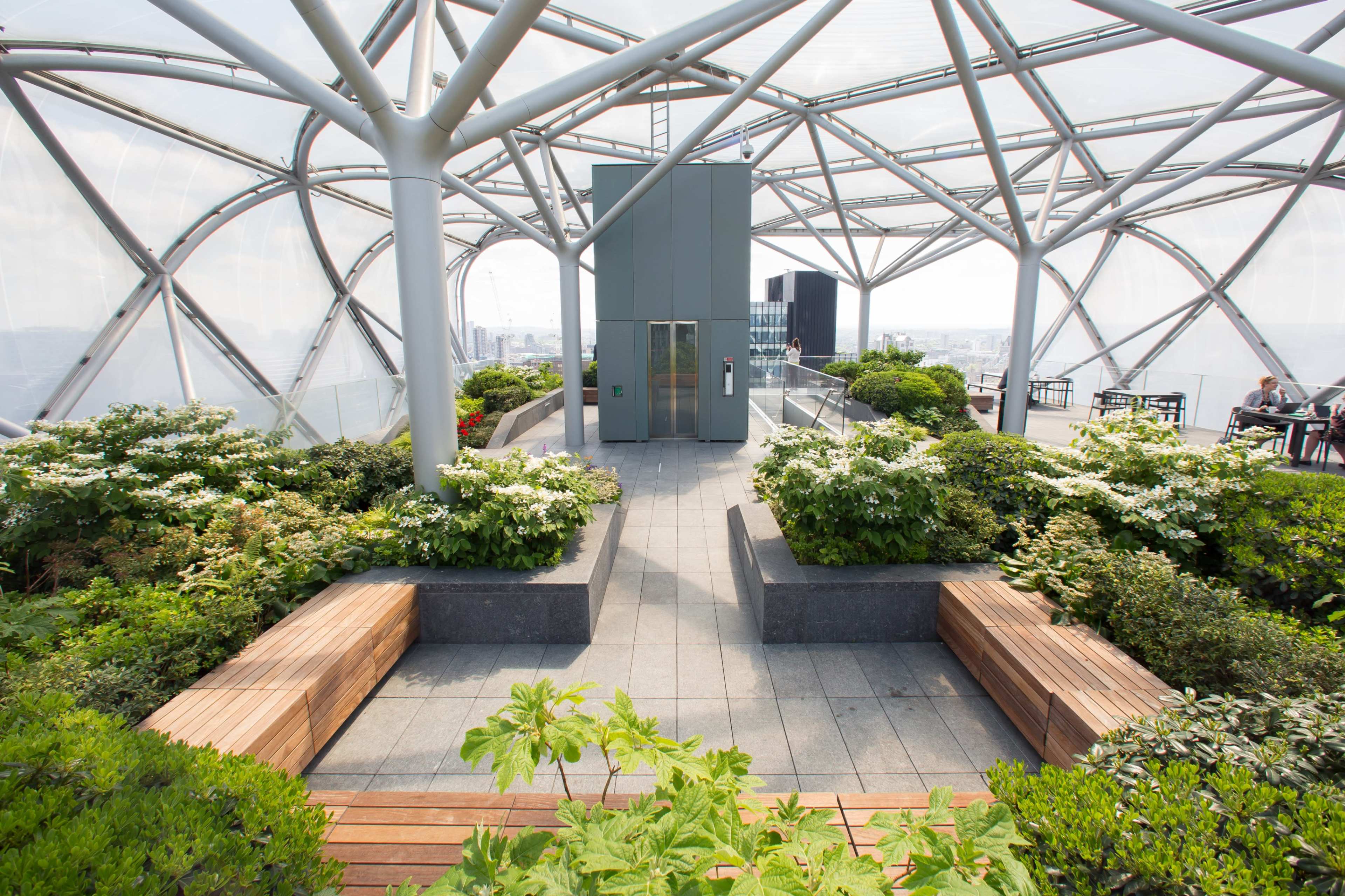 The image shows a rooftop garden terrace with a geometric glass structure, featuring green plants, wooden benches, and an elevator in the center.