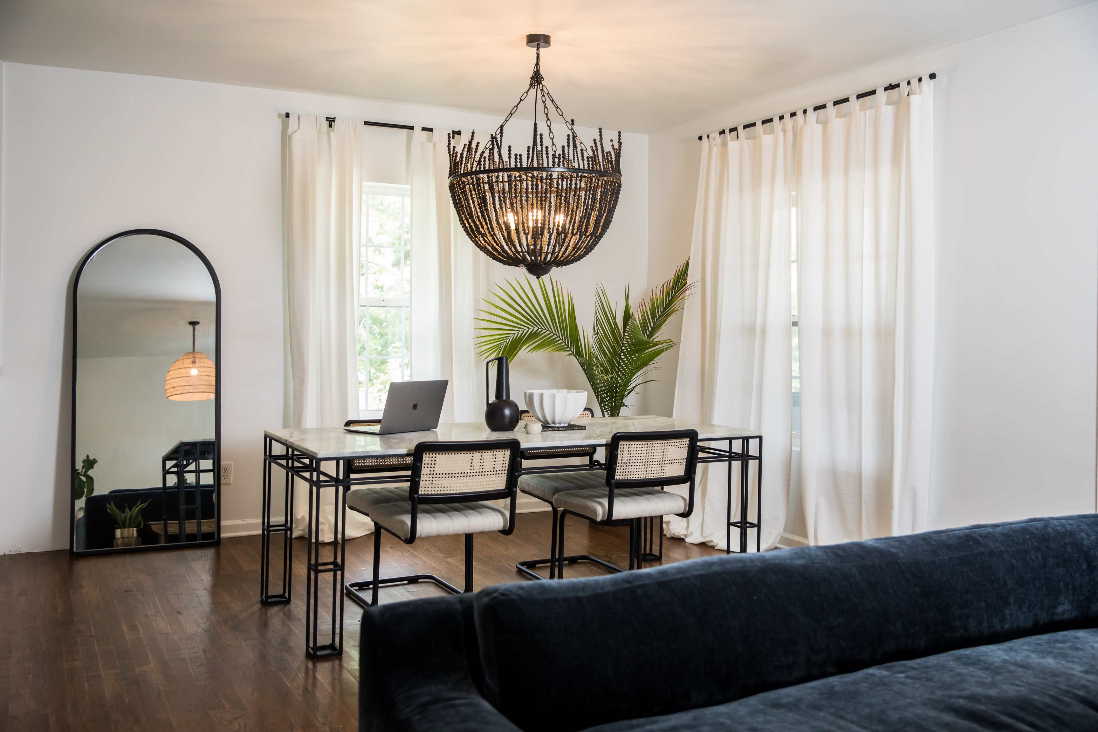 A dining area features a marble table with four chairs, a large mirror, and a stylish chandelier, surrounded by bright curtains and a potted plant.