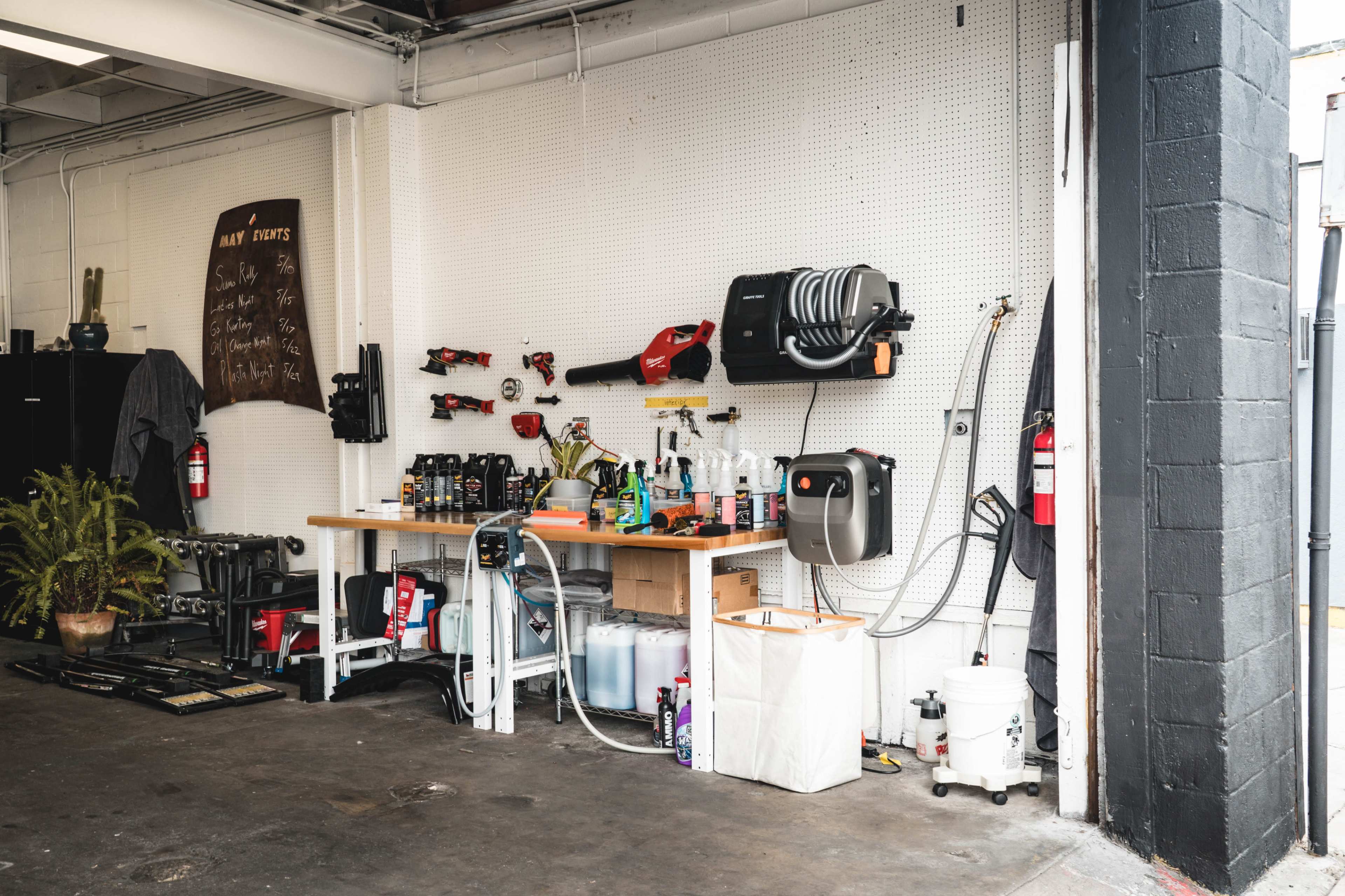 The image shows a workspace with a pegboard featuring various tools, a wooden workbench equipped with equipment and supplies, and a plant in the corner.