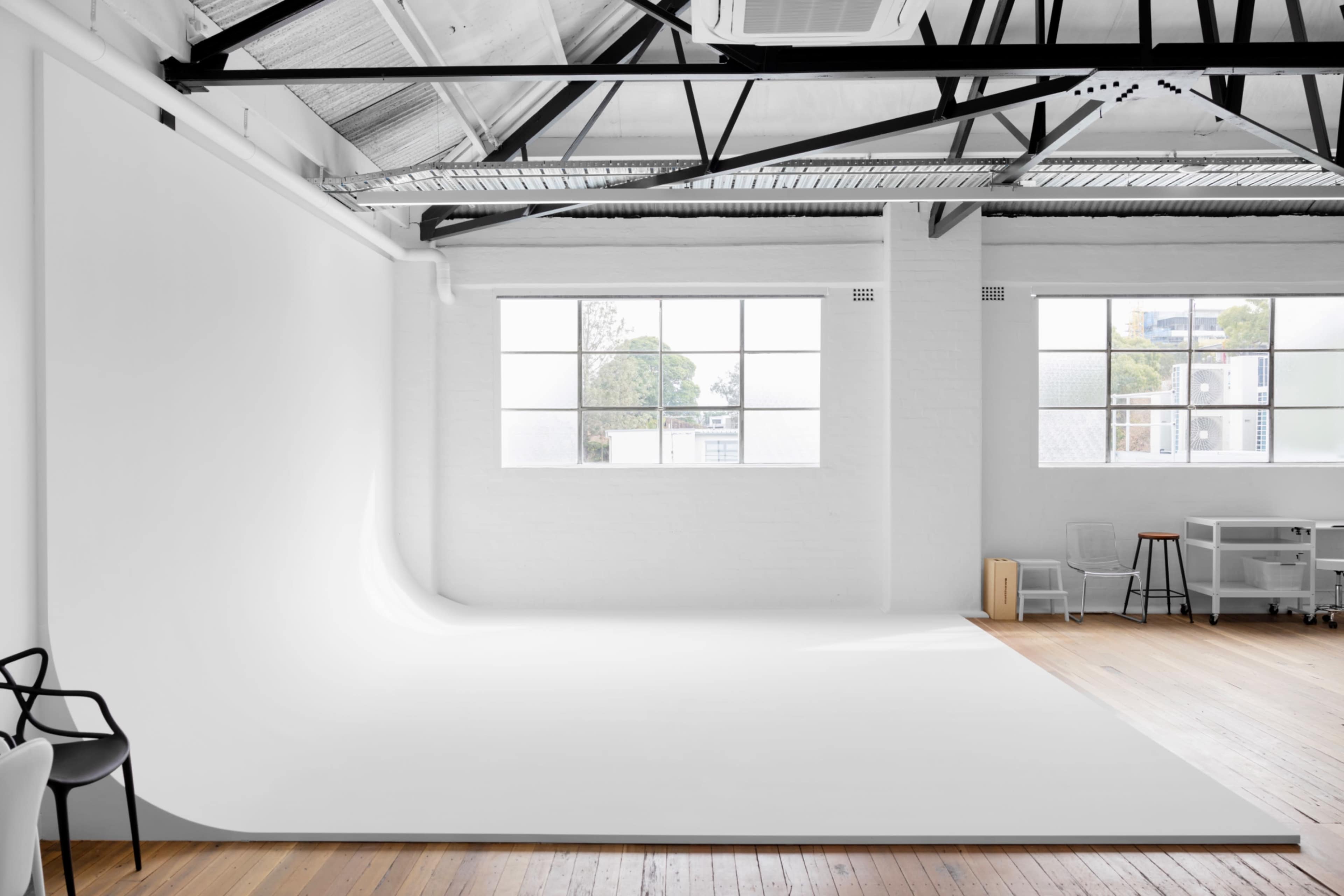 A spacious photography studio with white walls, a seamless backdrop, large windows, and wooden flooring.