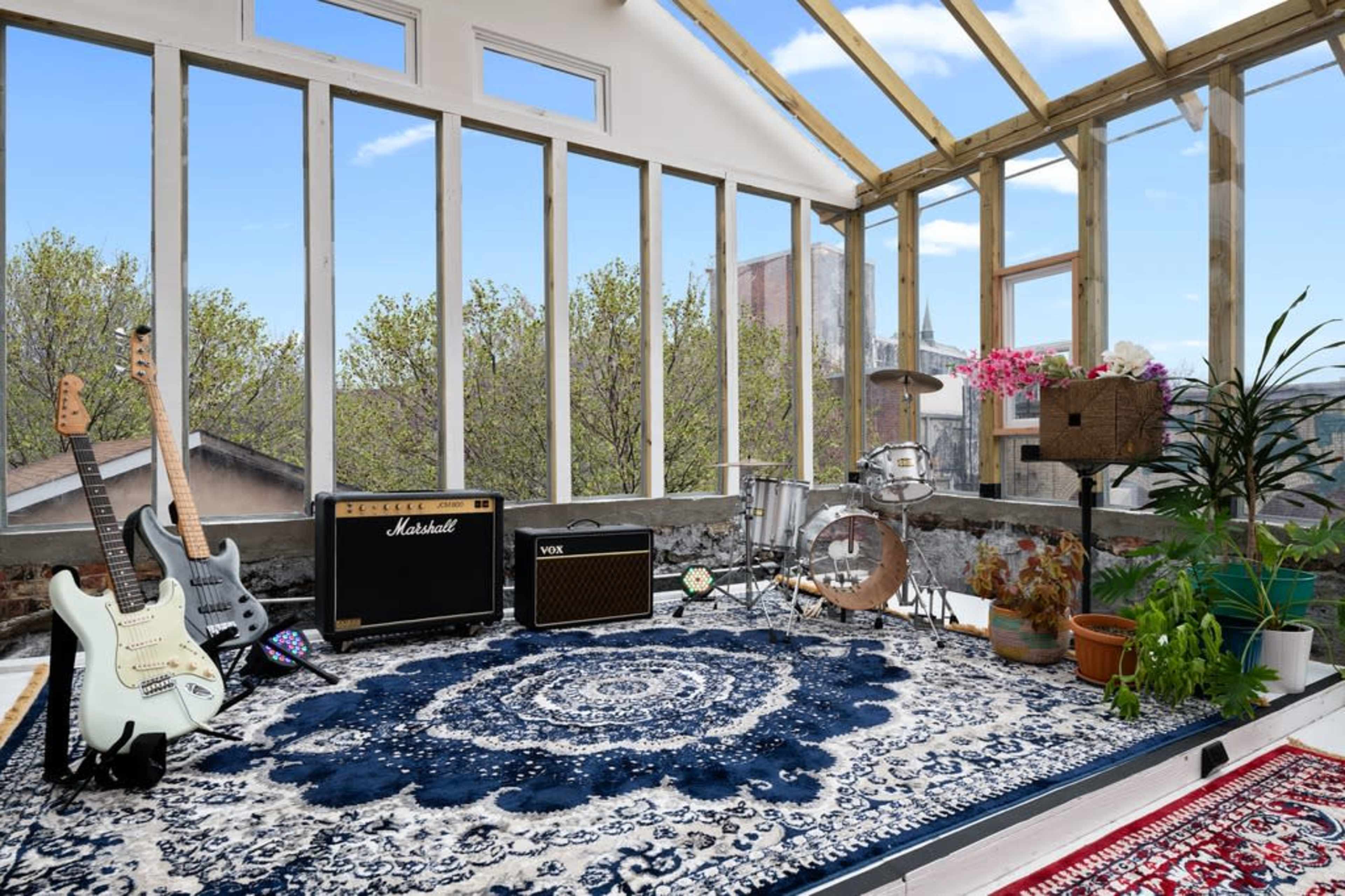 A sunlit music room features electric guitars, amplifiers, and a drum set arranged around a decorative rug, with large windows offering a view of trees outside.