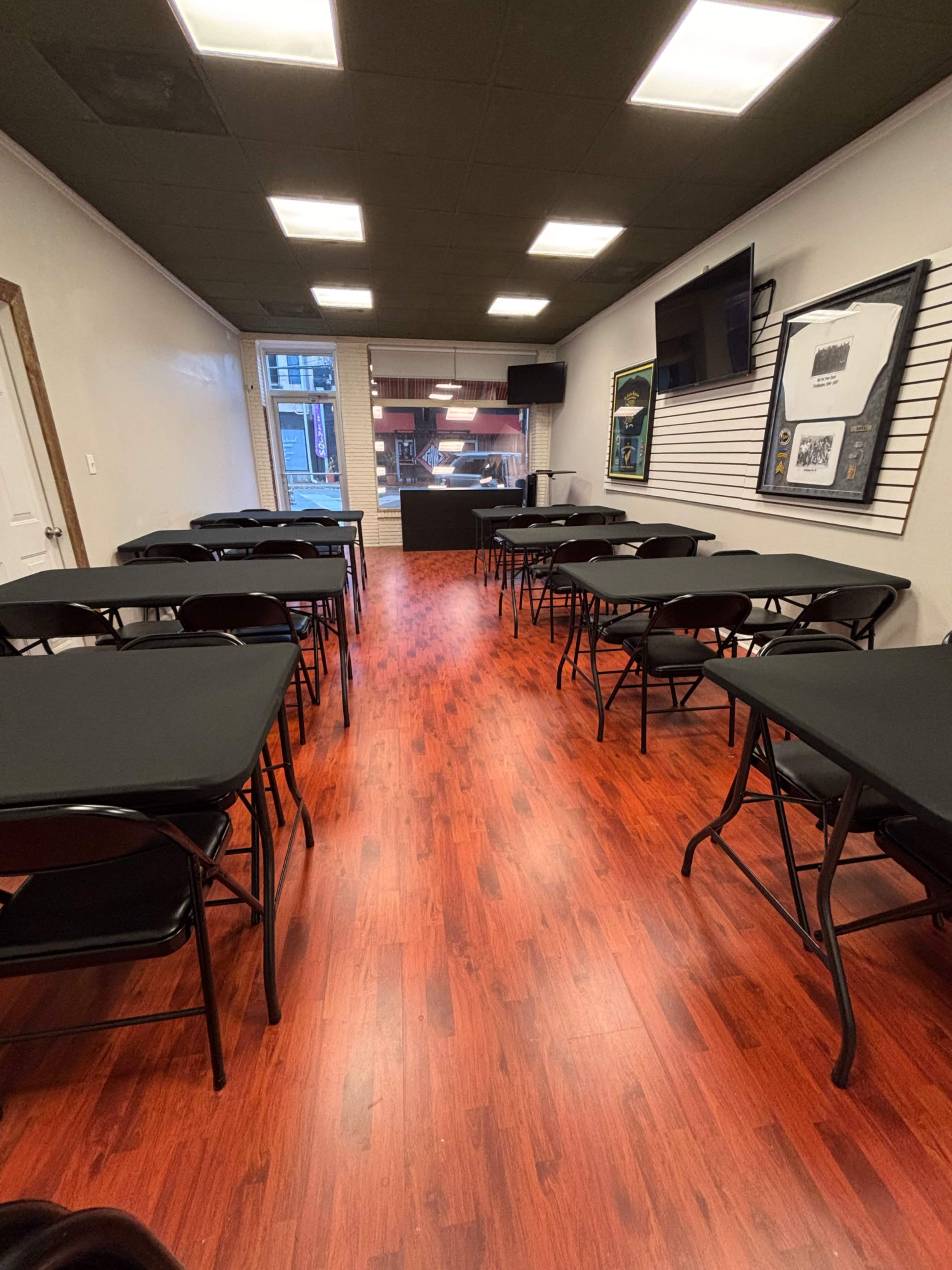 The image shows a spacious room with several rows of black folding tables and chairs set up around a central area, illuminated by overhead lights and featuring a large screen on one wall.