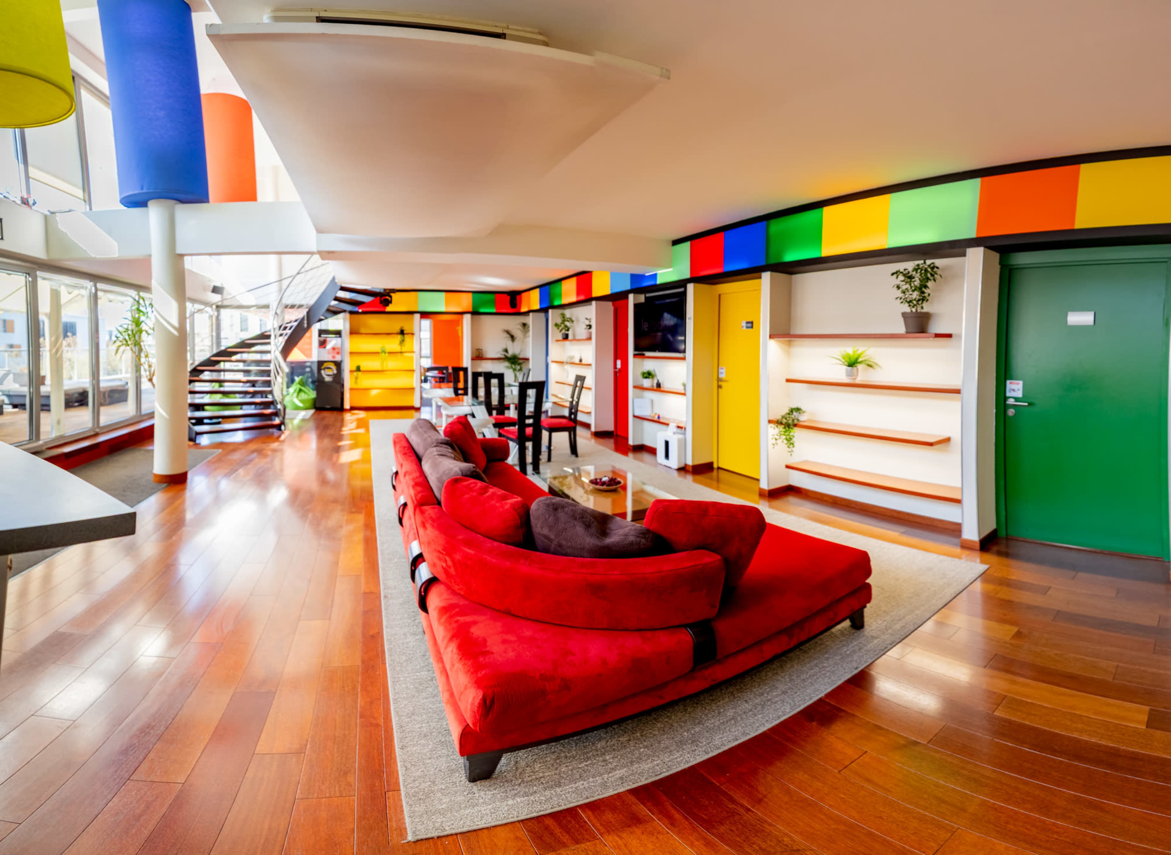 The image shows a brightly colored modern living space featuring a red sofa set, a dining area, and a staircase in a well-lit, open layout.