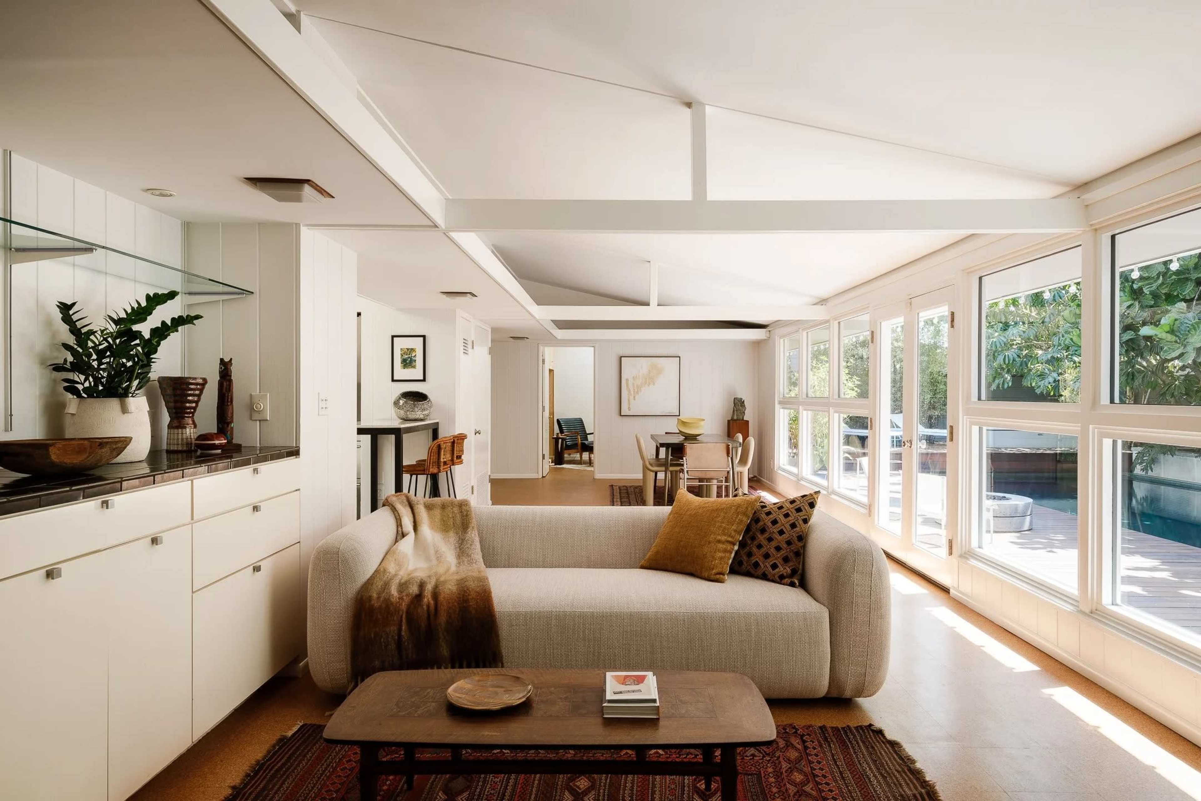 The image shows a modern living room with a light-colored sofa, a wooden coffee table, and large windows that allow natural light to fill the space.