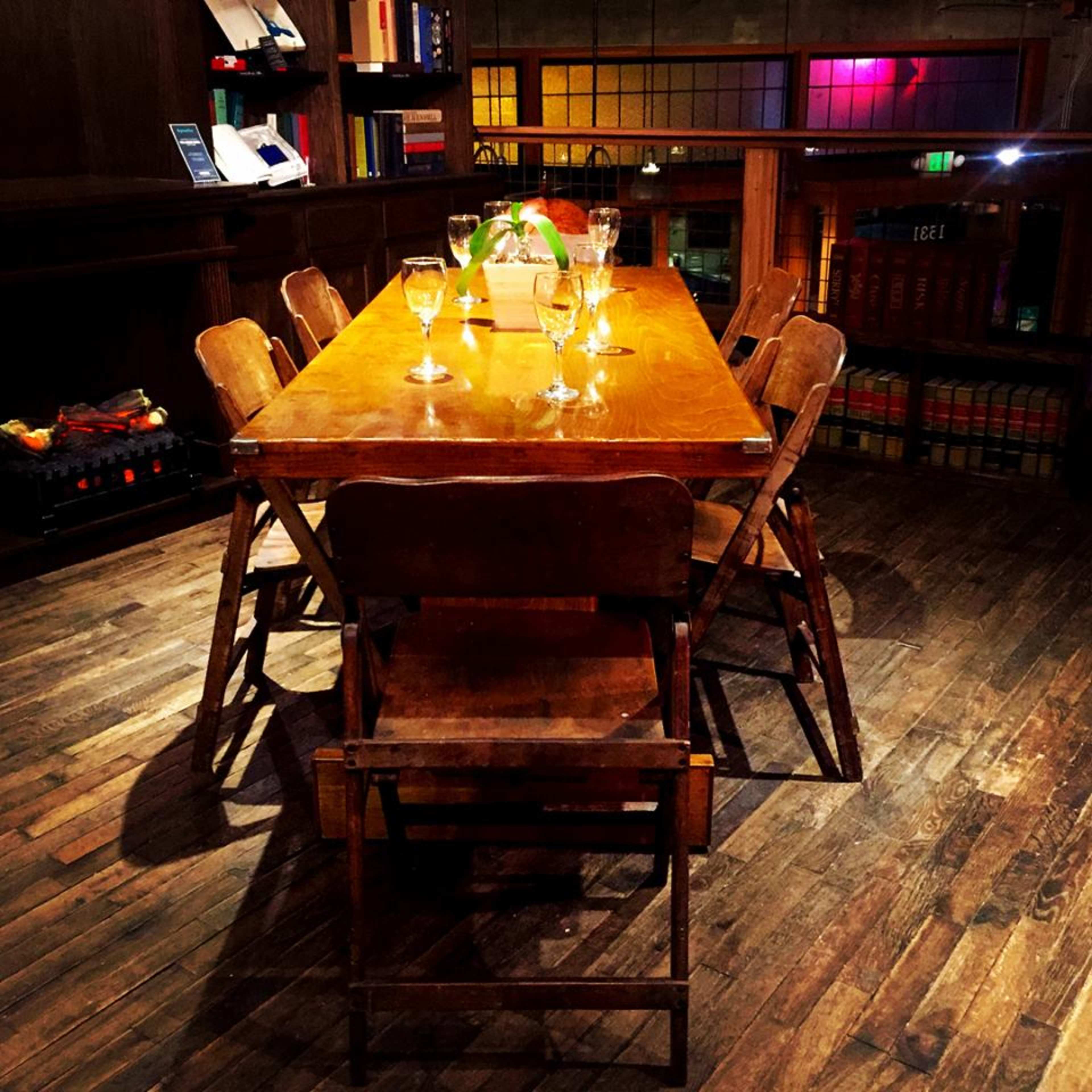 A wooden dining table is set with empty glassware, surrounded by wooden chairs in a dimly lit room with a bookshelf and colorful lights in the background.