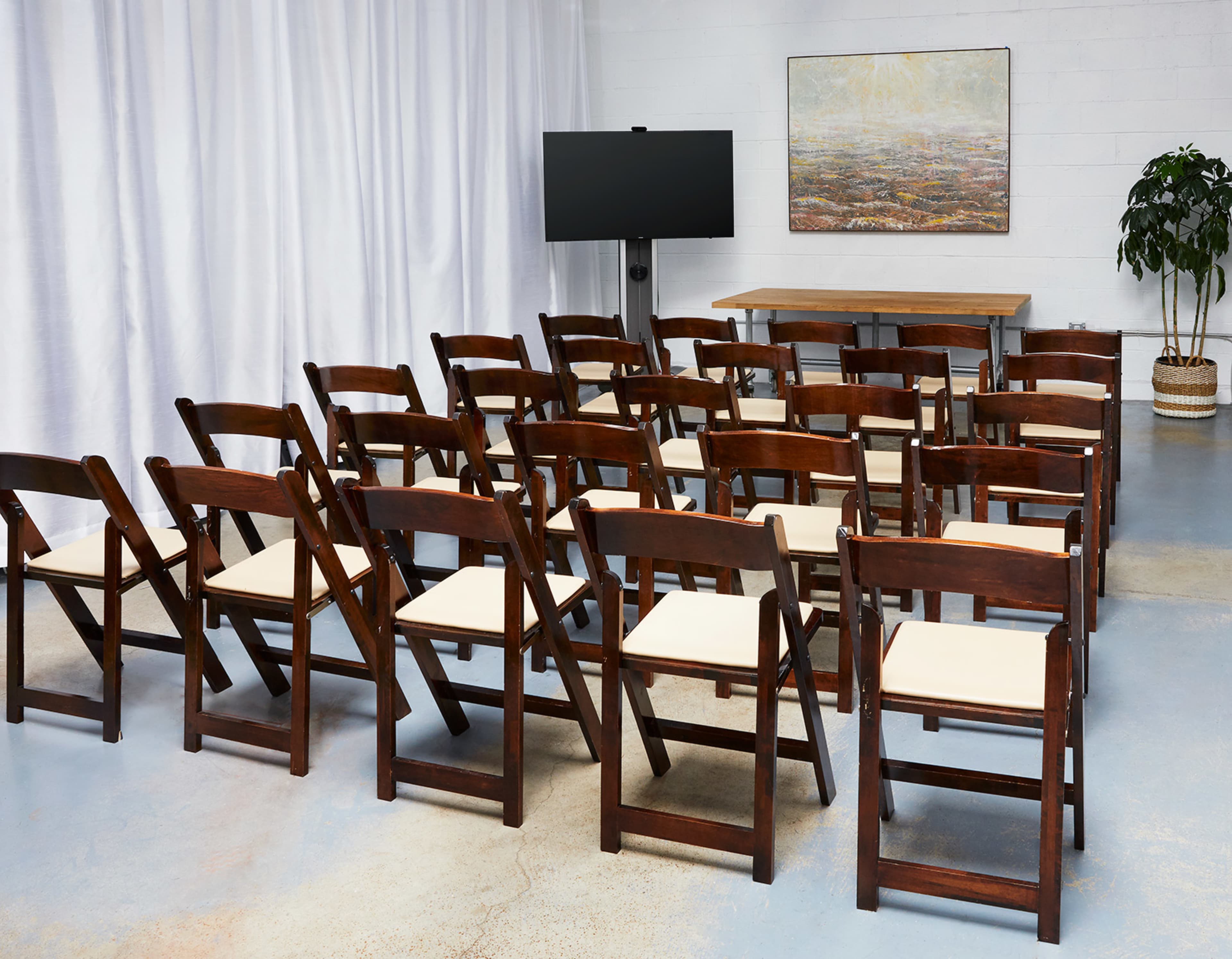A set of wooden folding chairs is arranged in rows facing a display screen in a room with white drapes and a potted plant.