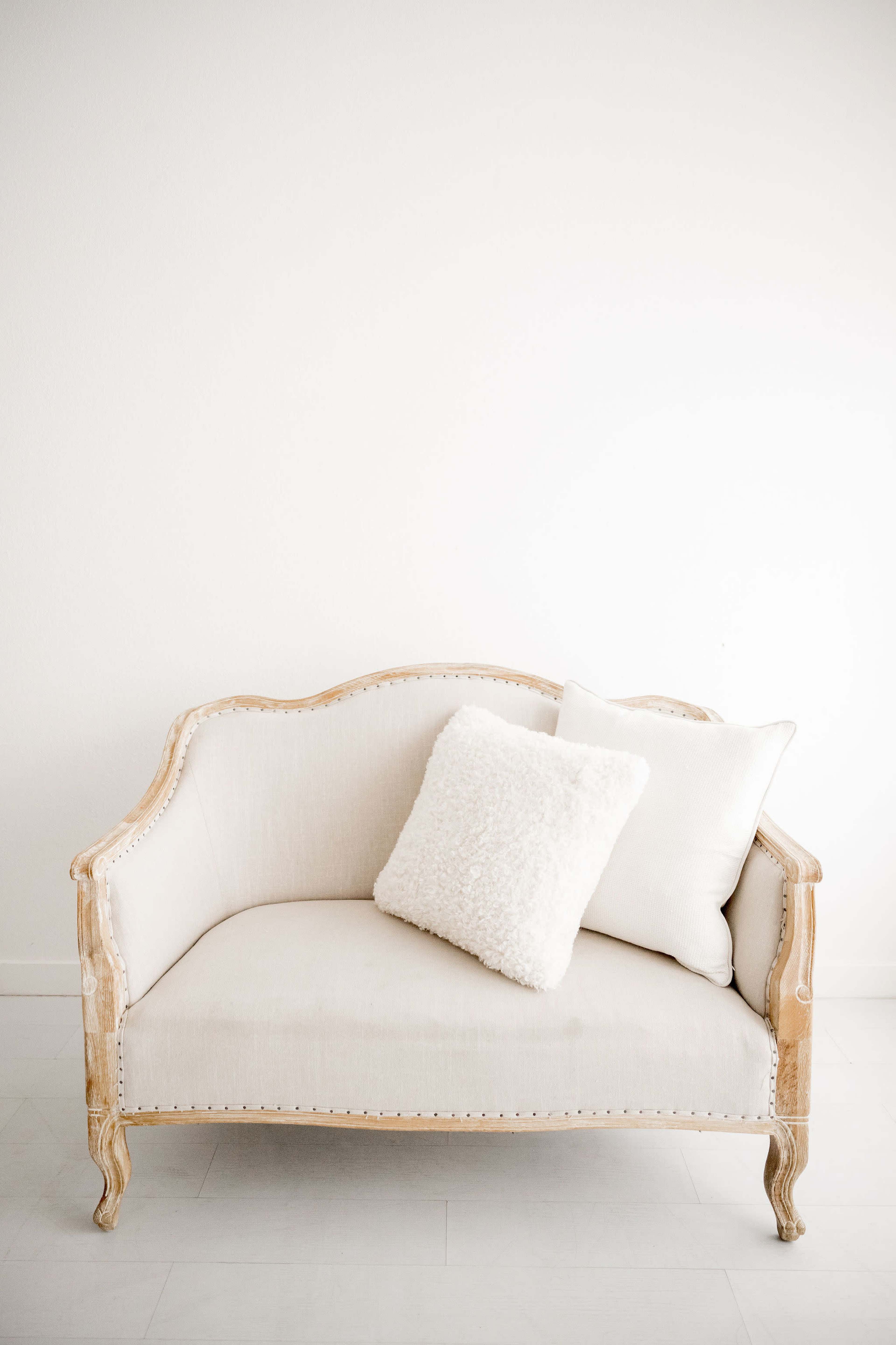 A light-colored vintage couch with decorative carvings and two pillows sits against a plain white wall.