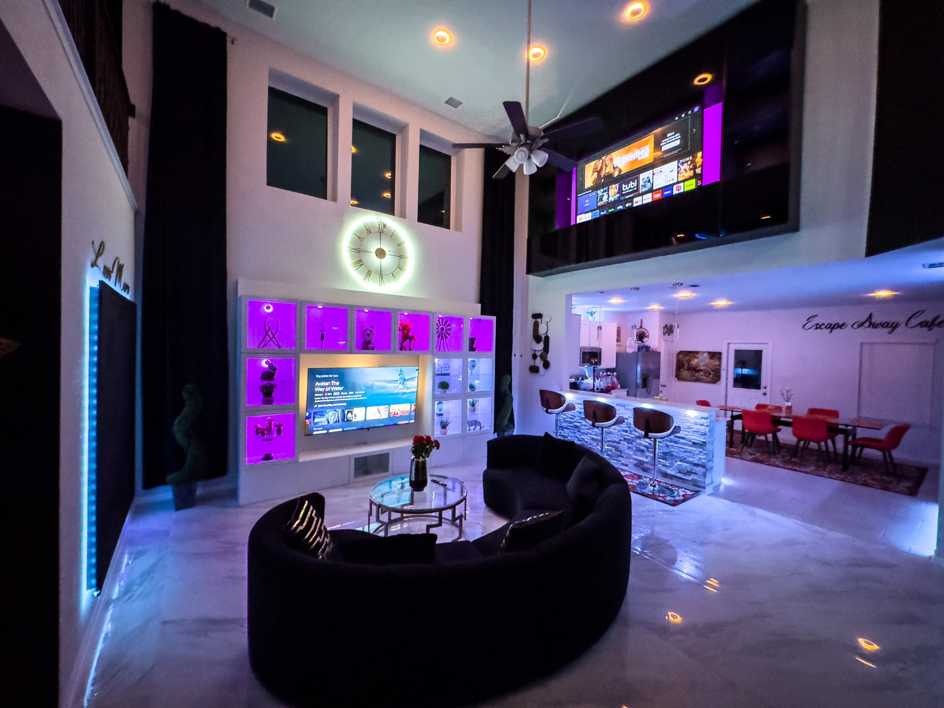 A modern living room features a curved black sofa, illuminated shelves, and a large wall-mounted TV, with bright purple and white lighting accents.