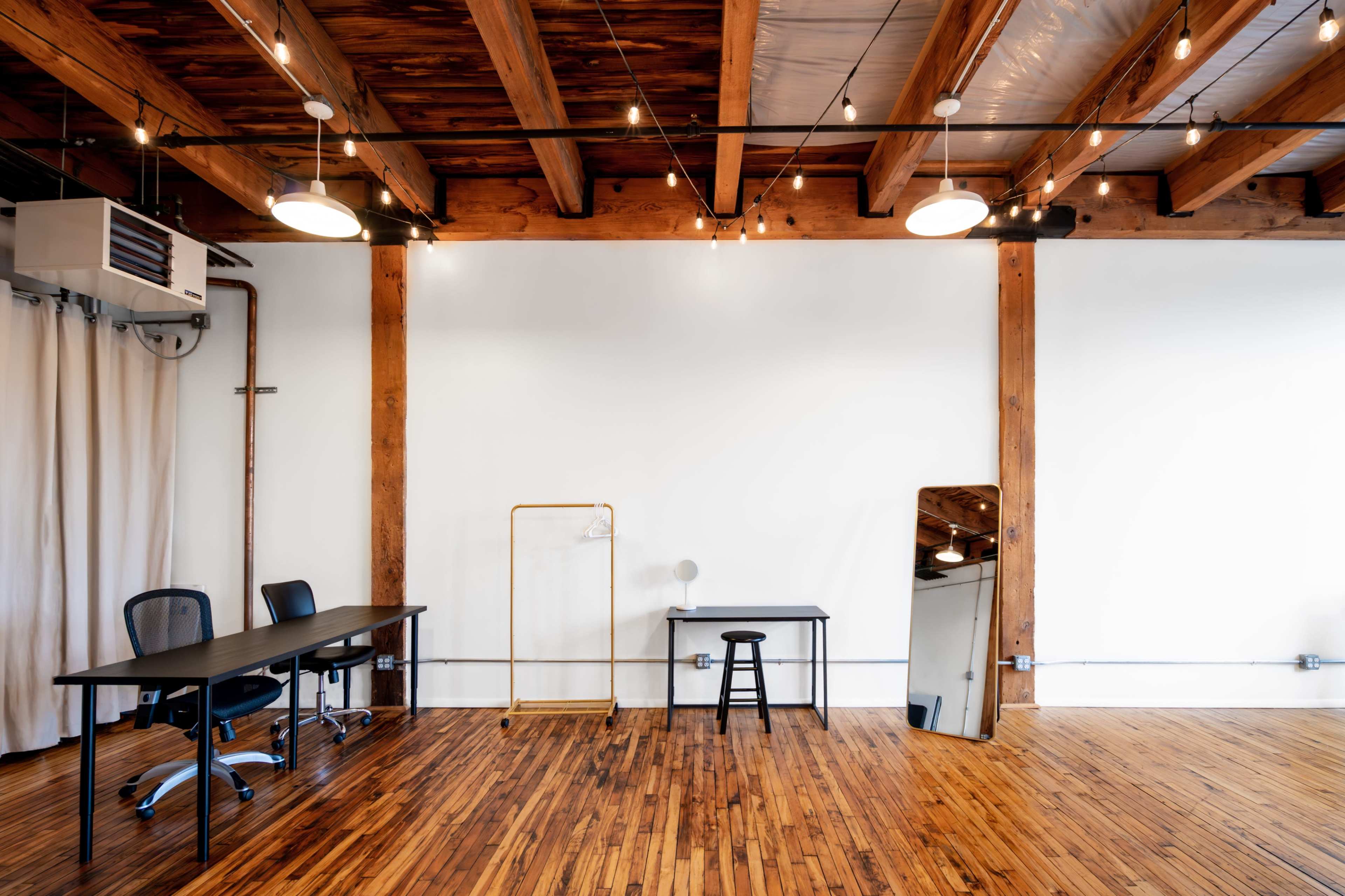 A spacious, minimalist office layout with wooden floors, exposed beams, and a few pieces of furniture including two desks, a stool, and a mirror.