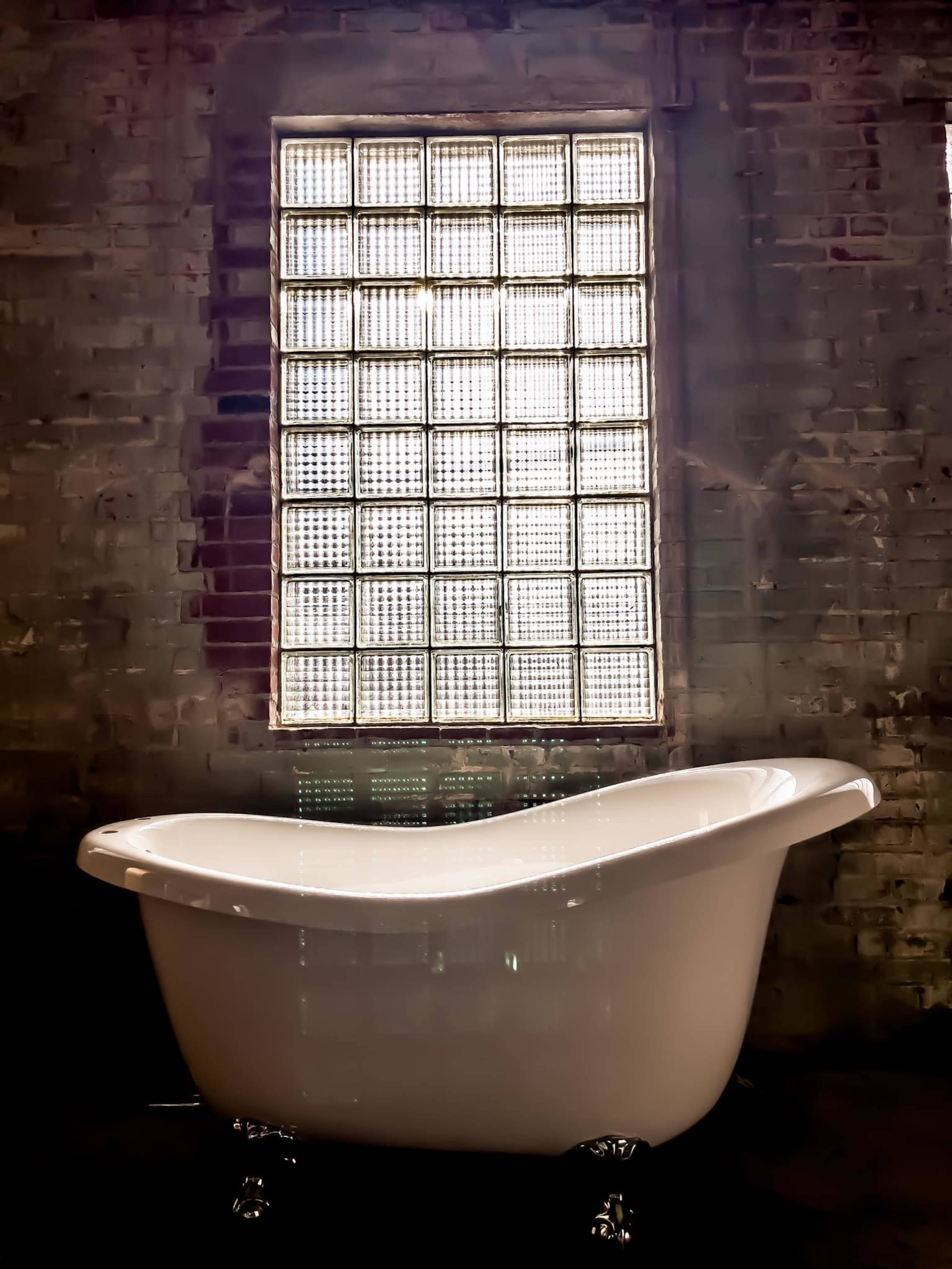 A white freestanding bathtub is positioned in front of a large glass block window set into a brick wall.