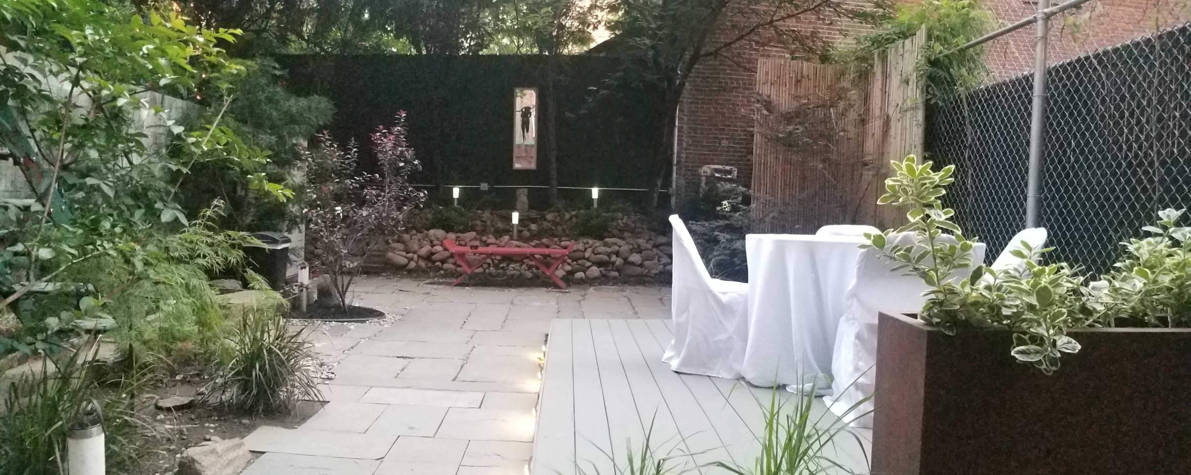 A small outdoor garden area features a stone path, a wooden bench, and a table covered with a white tablecloth, surrounded by greenery and decorative rocks.