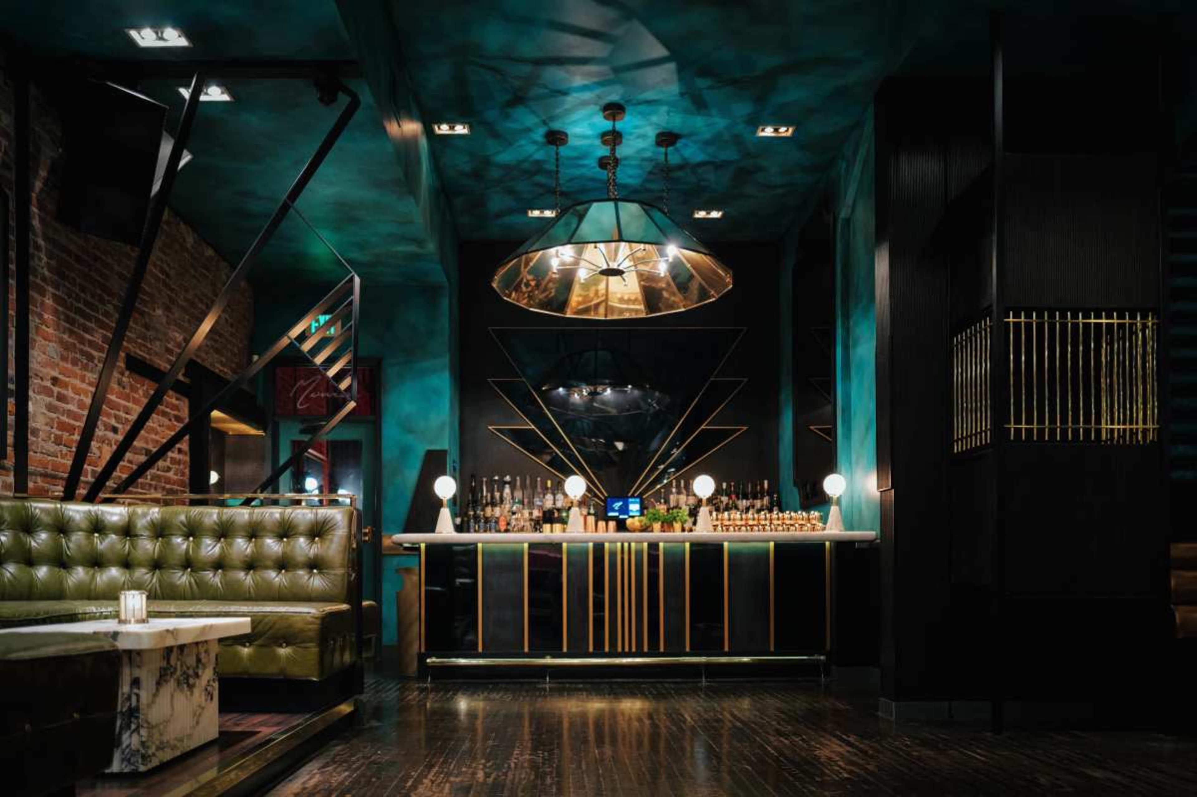 The image features a stylish bar interior with teal walls, a mirrored backdrop, and a dark wooden floor, complemented by a green velvet sofa and modern light fixtures.