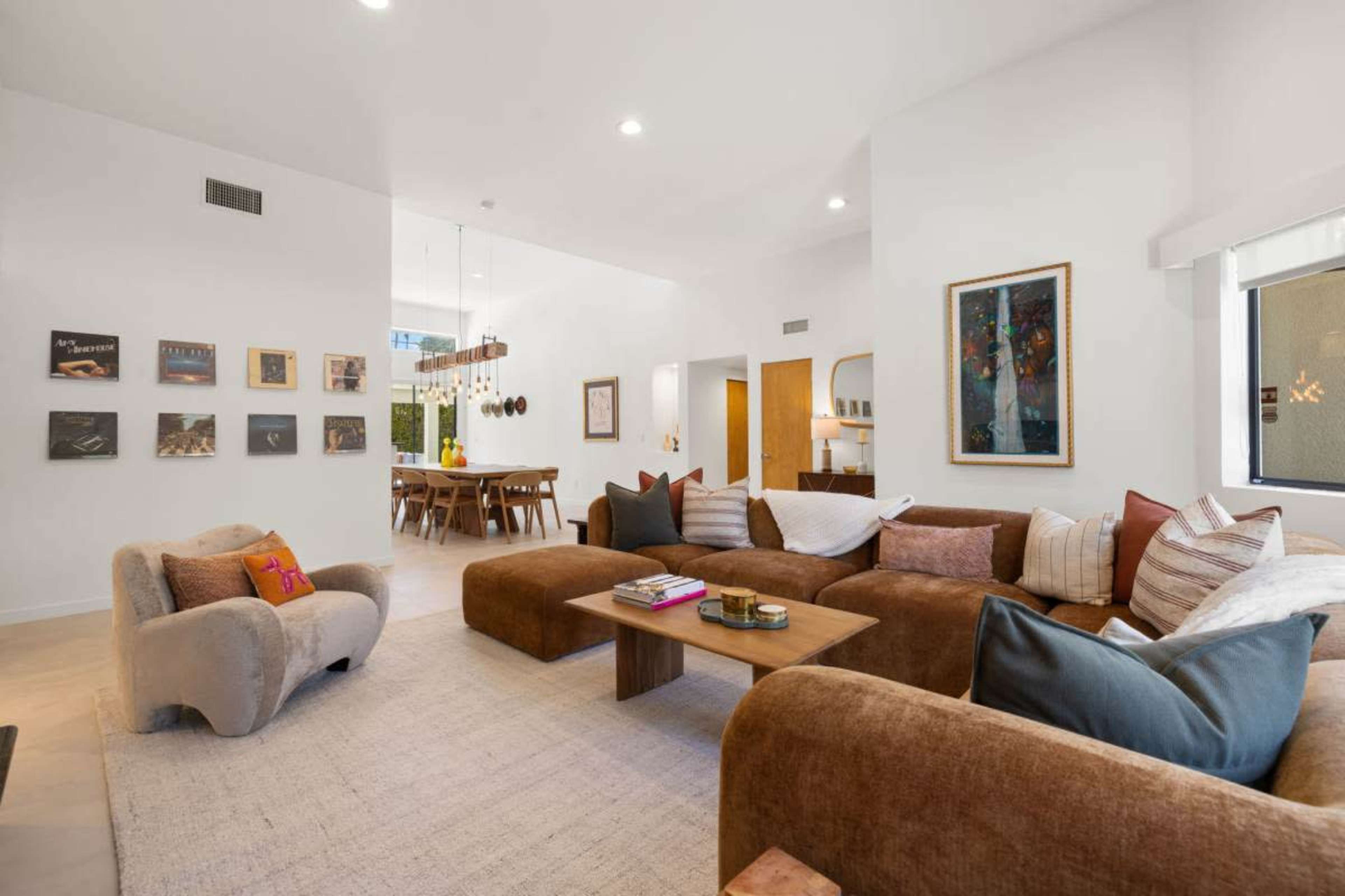 The image shows a modern living room featuring a large, brown sectional sofa, a wooden coffee table, and an arrangement of decorative pillows, with artwork displayed on the walls and a dining area visible in the background.