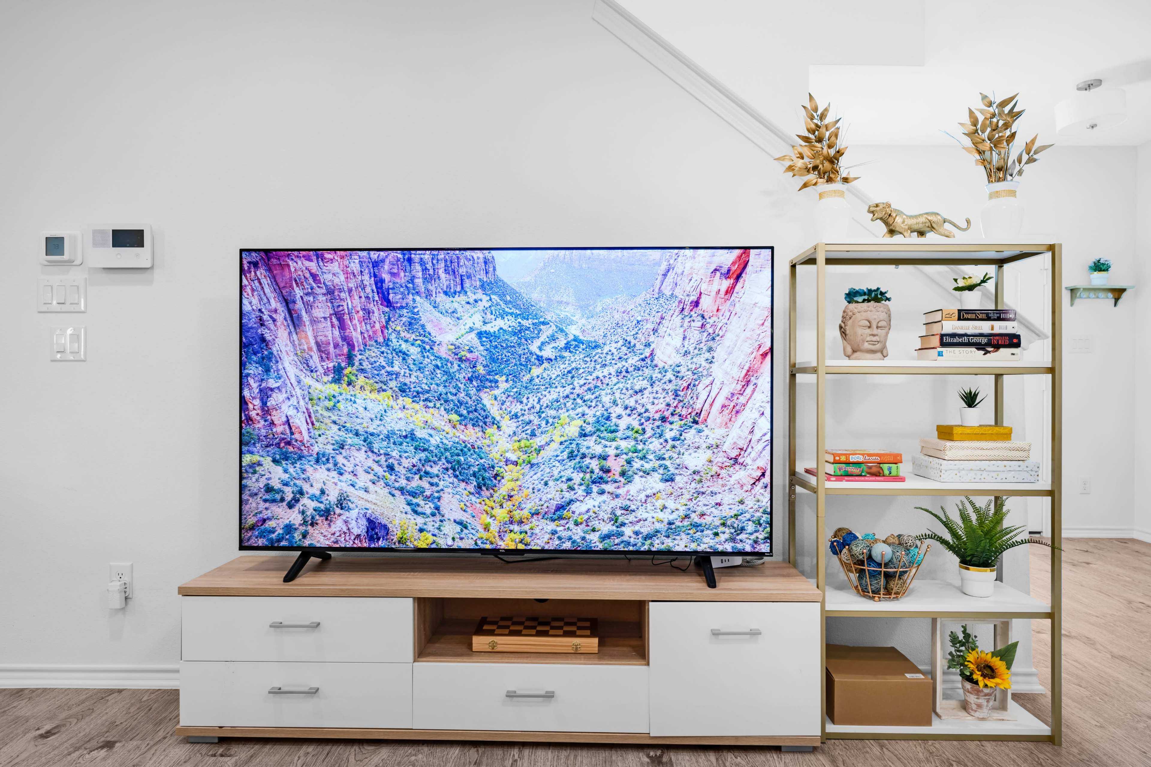 A large television displaying a mountainous landscape is positioned on a wooden console beside a shelf filled with decorations and books.