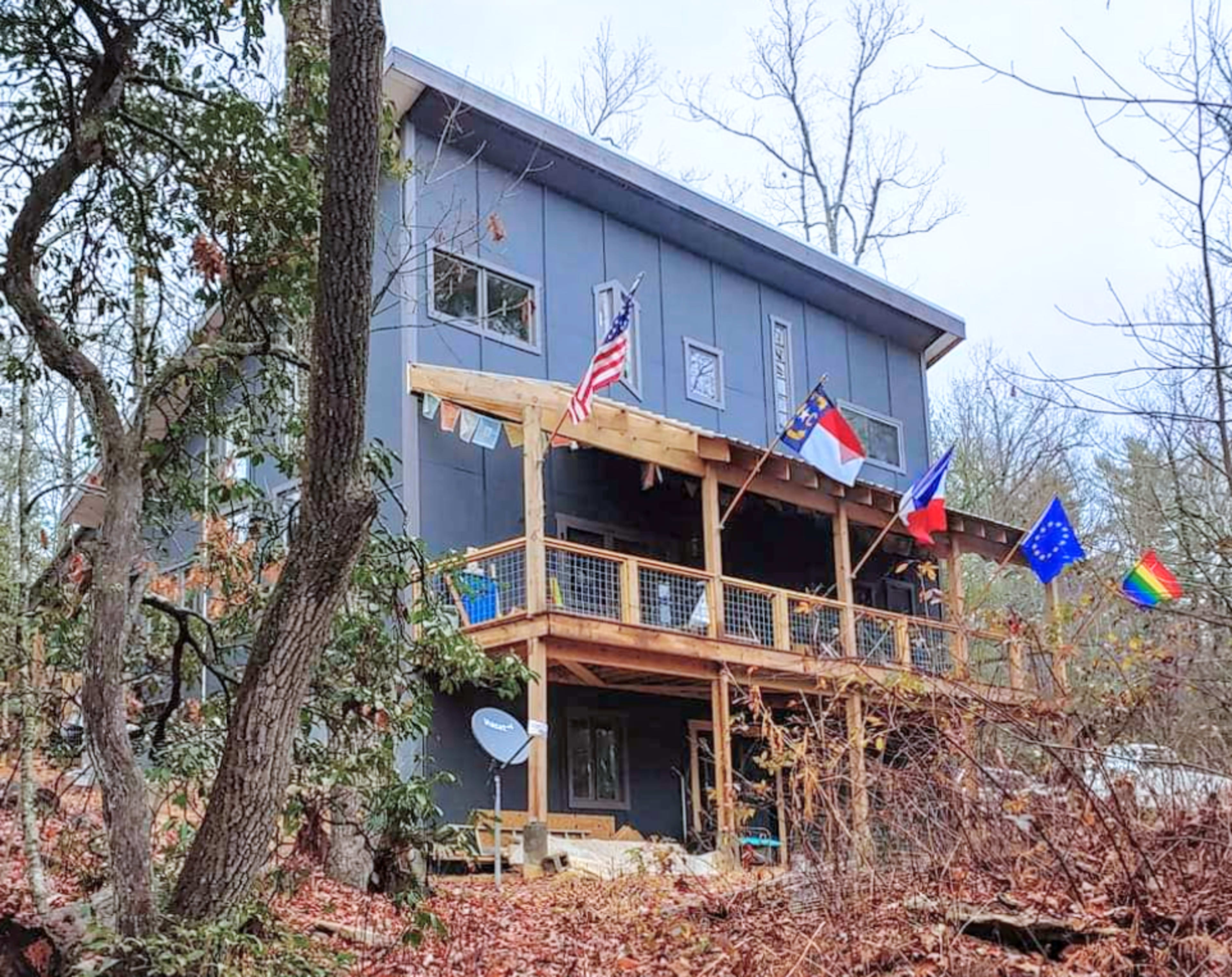 A three-story house with a wooden deck displays multiple flags, including the American flag, a state flag, and an EU flag, surrounded by autumn foliage.