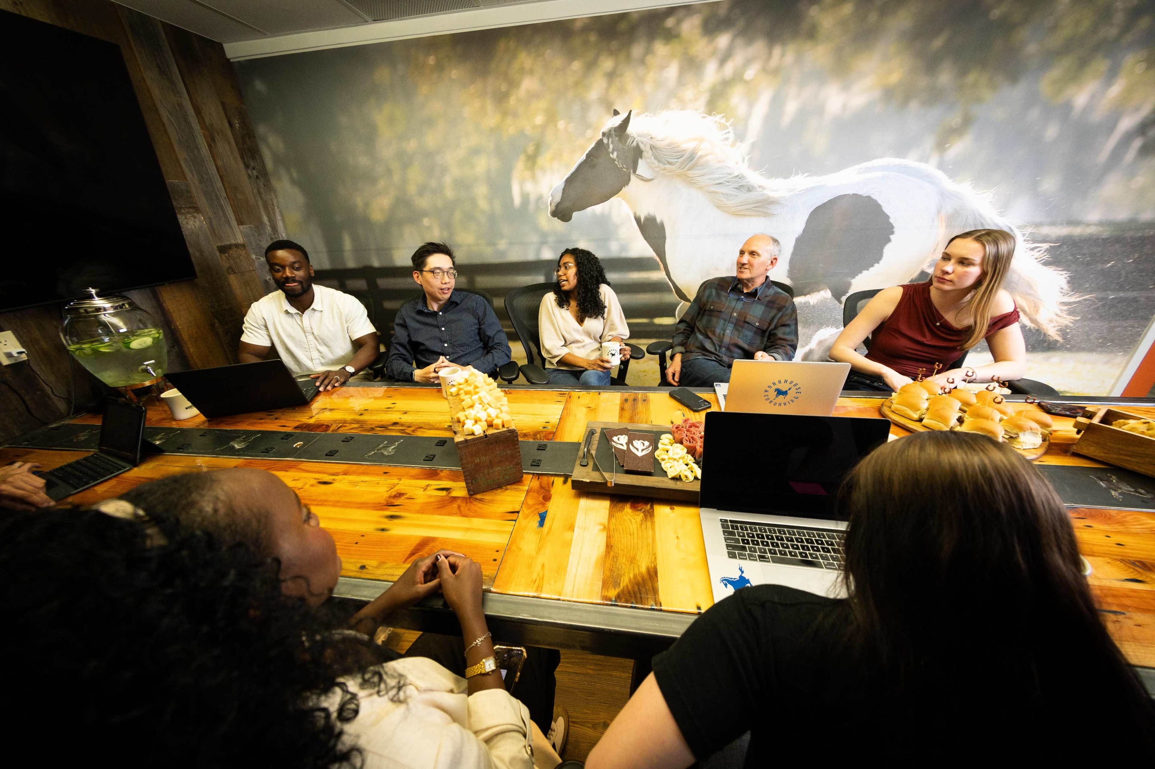 A group of six people is seated around a conference table in a meeting room with a horse mural on the wall.