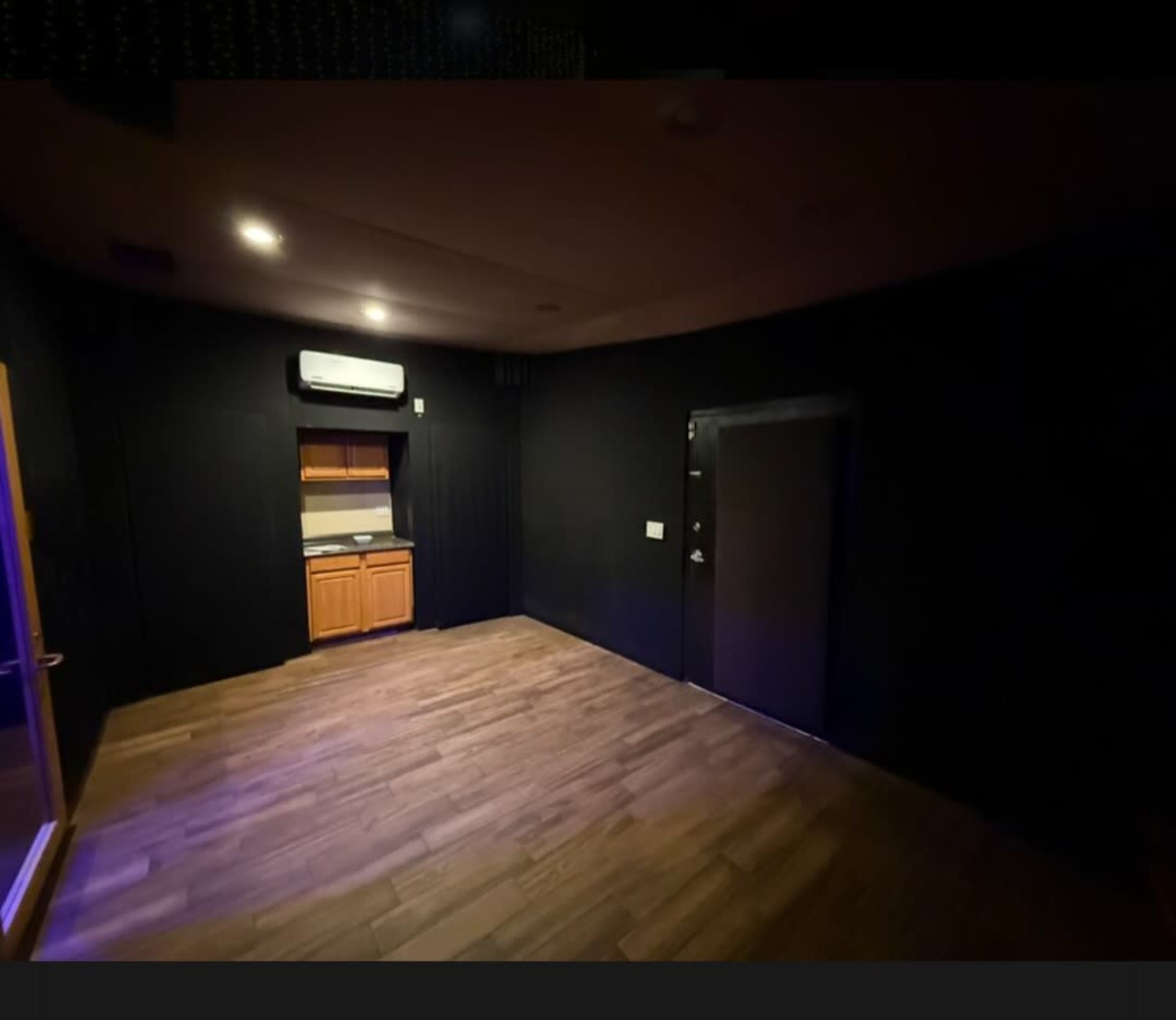The image shows a dimly lit room with black walls, a wooden floor, and a small kitchenette in one corner.