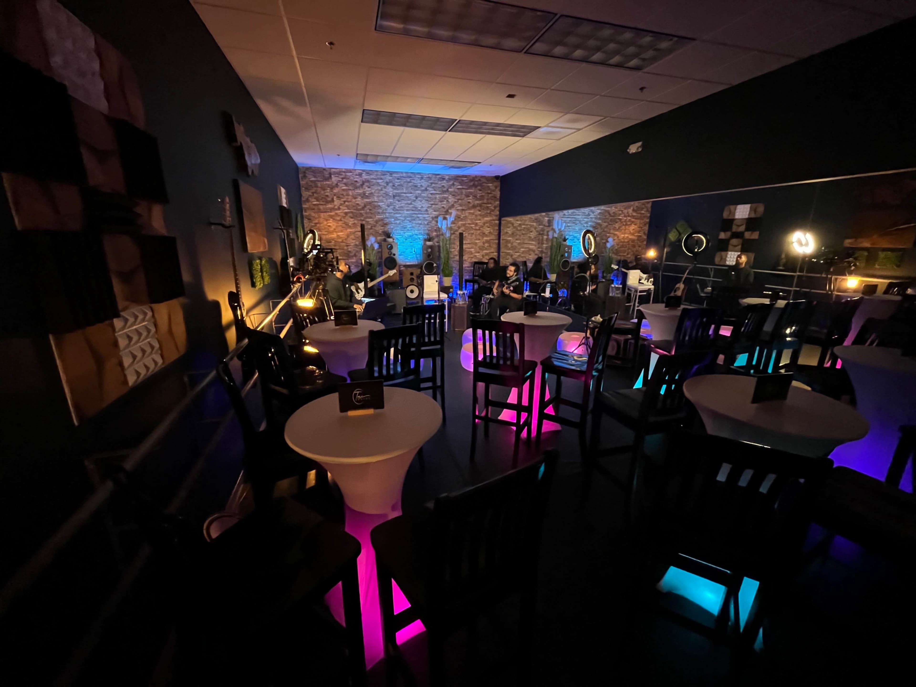 A dimly lit lounge with white tables and black chairs arranged in rows, featuring colorful lighting and decorative walls.