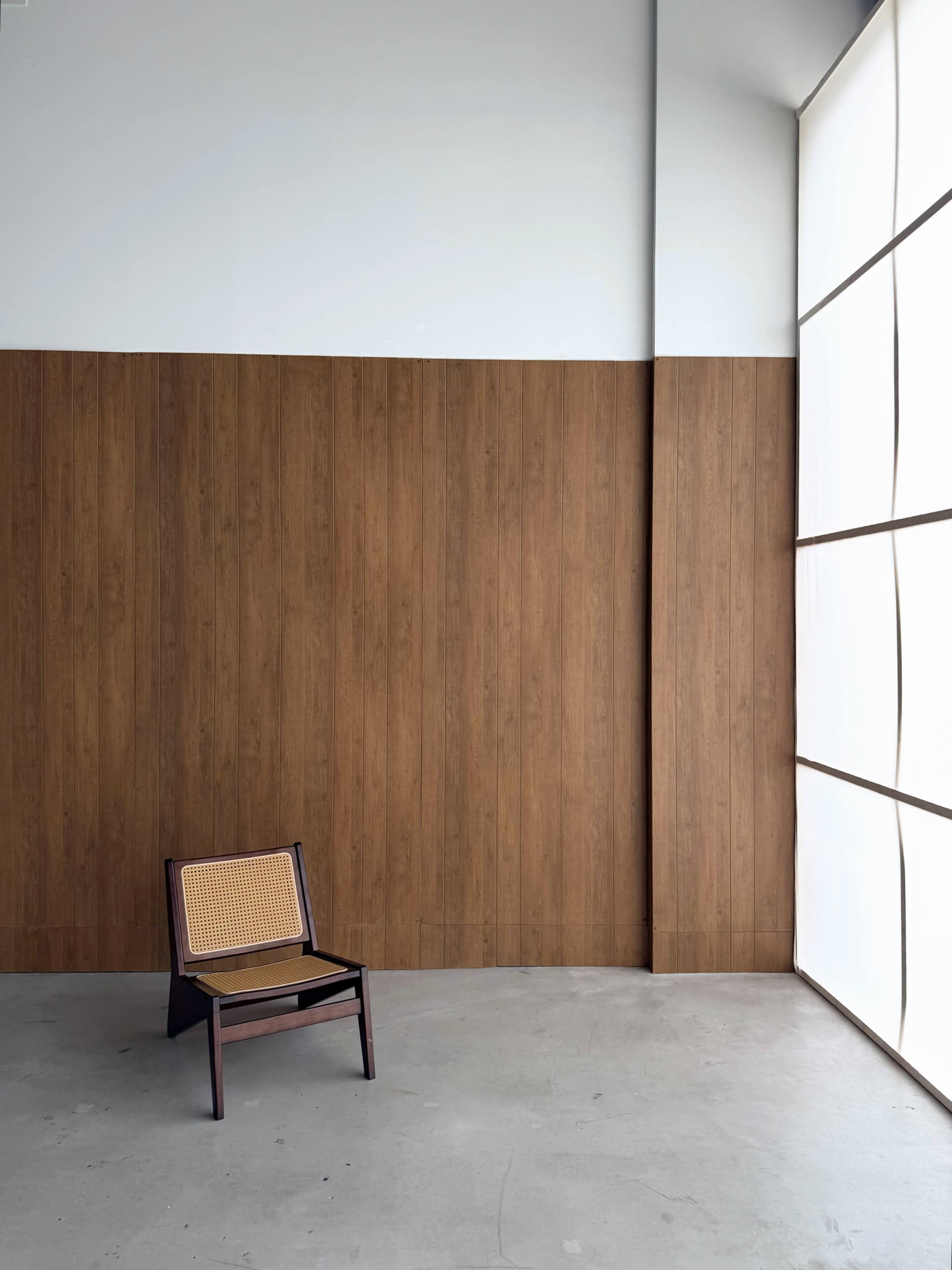 A wooden chair with a woven seat is positioned on a concrete floor in front of a large window and a wooden-paneled wall.