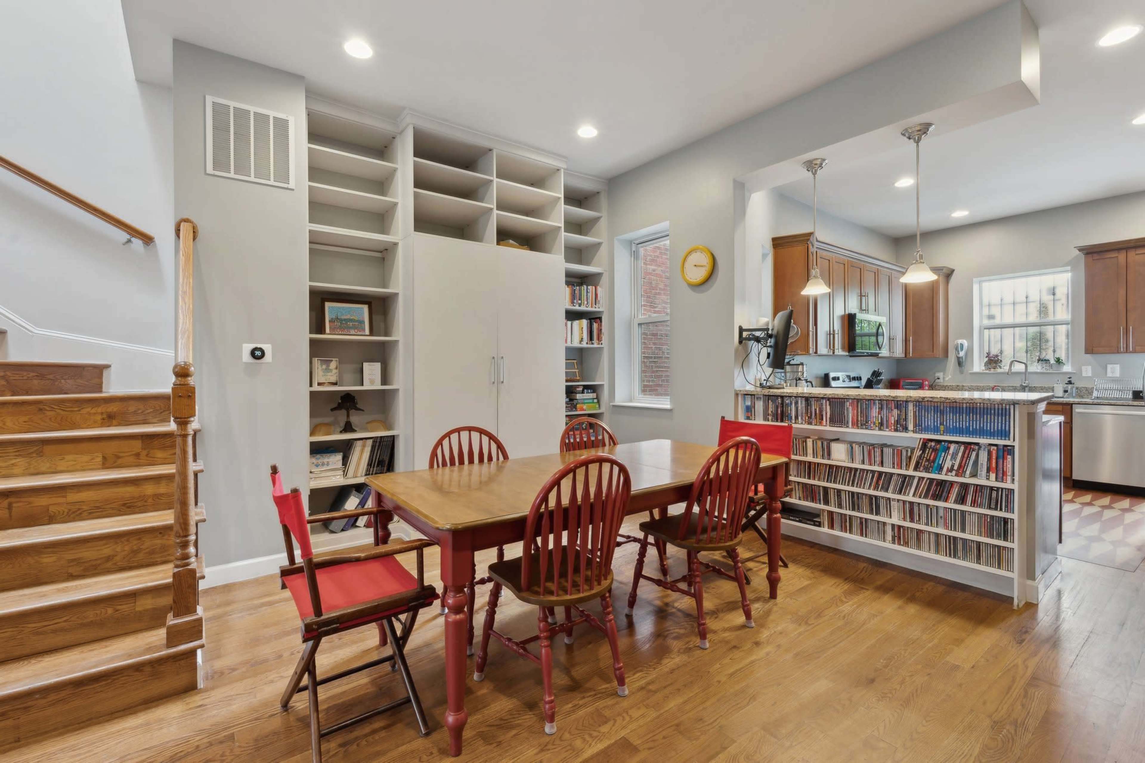 A dining area with a red table and chairs is situated next to a set of wooden stairs and a large bookshelf in a modern home.