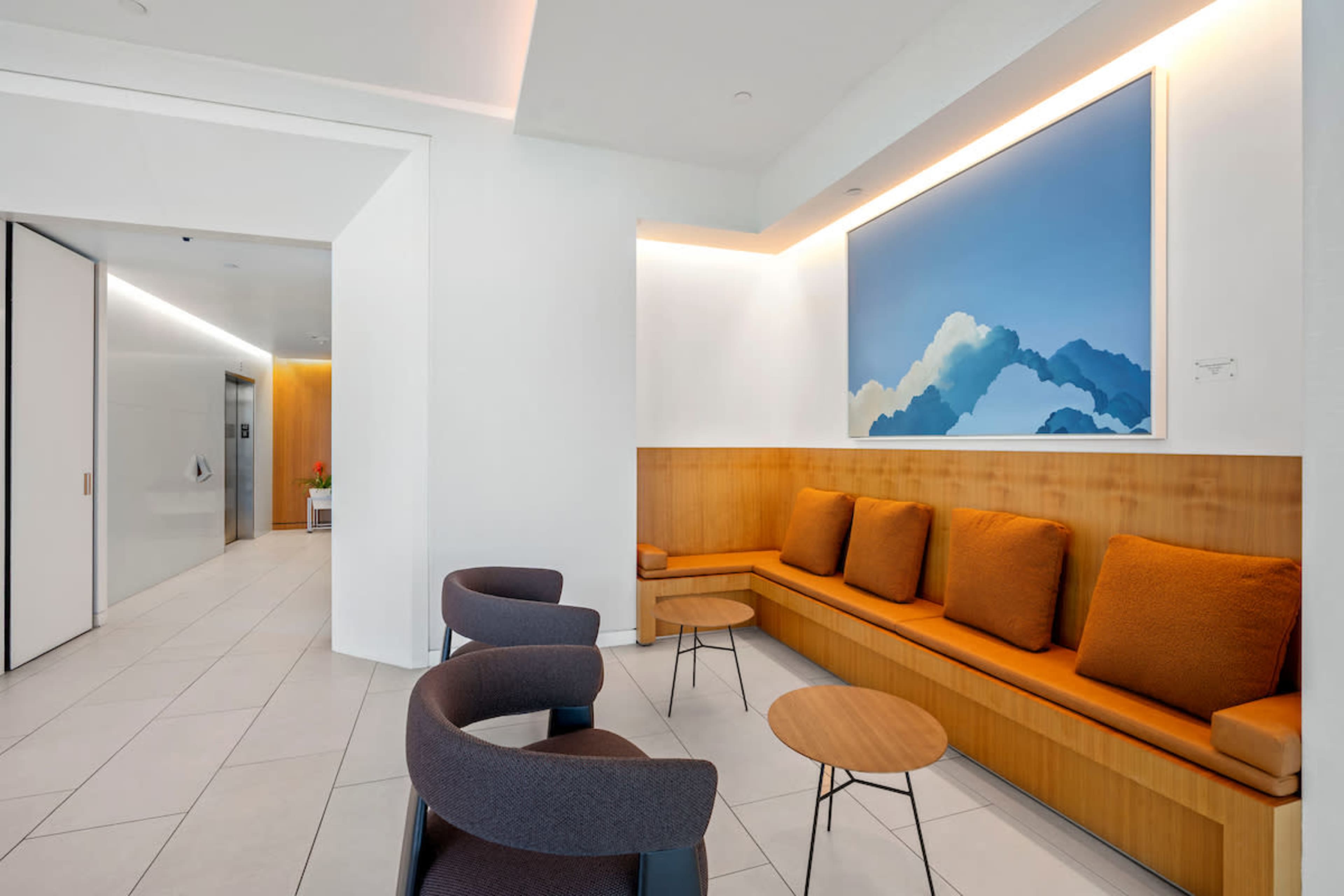 The image shows a modern waiting area with a wooden bench and round tables, accompanied by gray and orange chairs, and a large artwork featuring clouds on the wall.