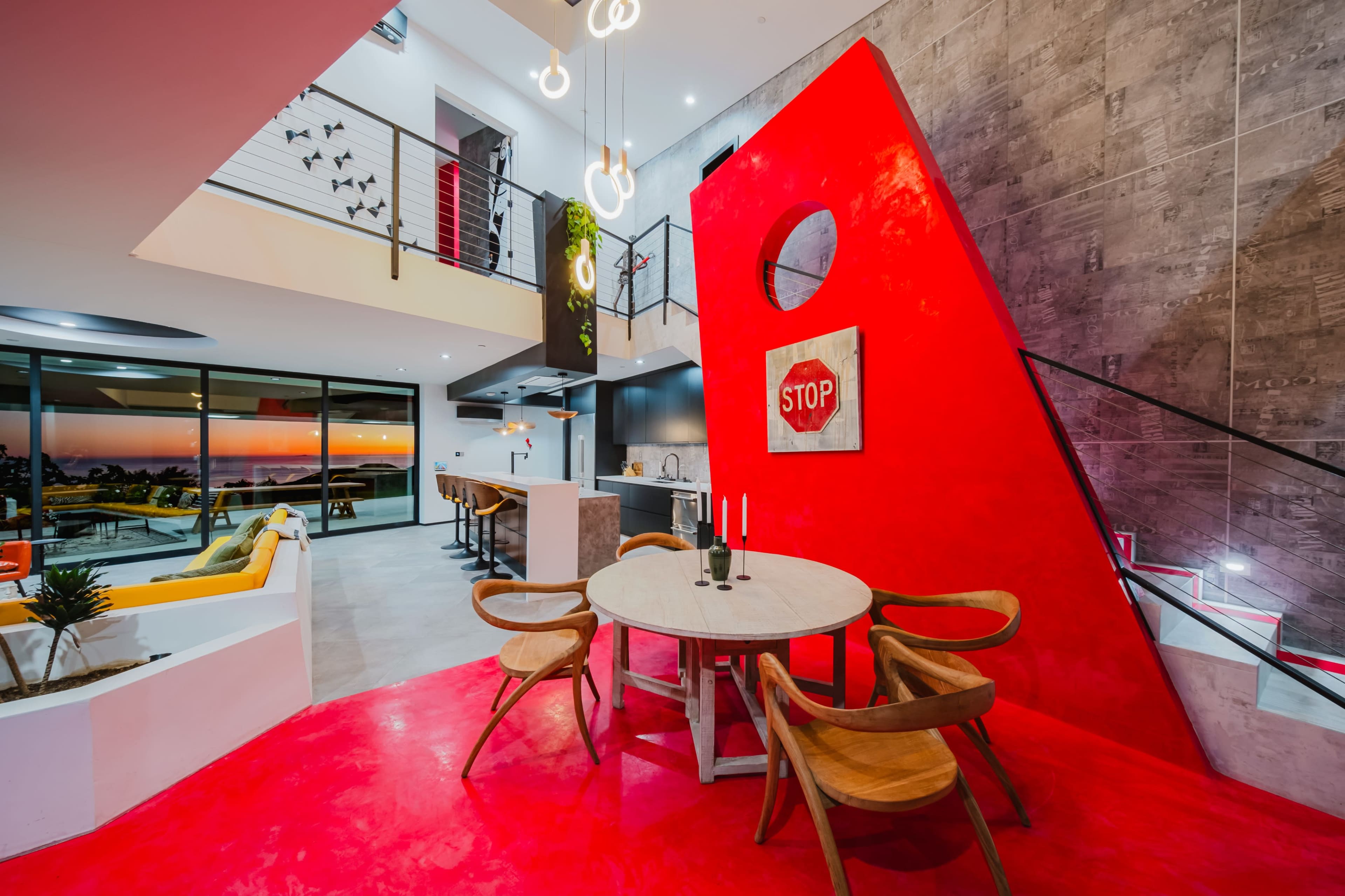 The image shows a modern living space with a red accent wall, a round dining table with four wooden chairs, and a staircase leading to an upper level.