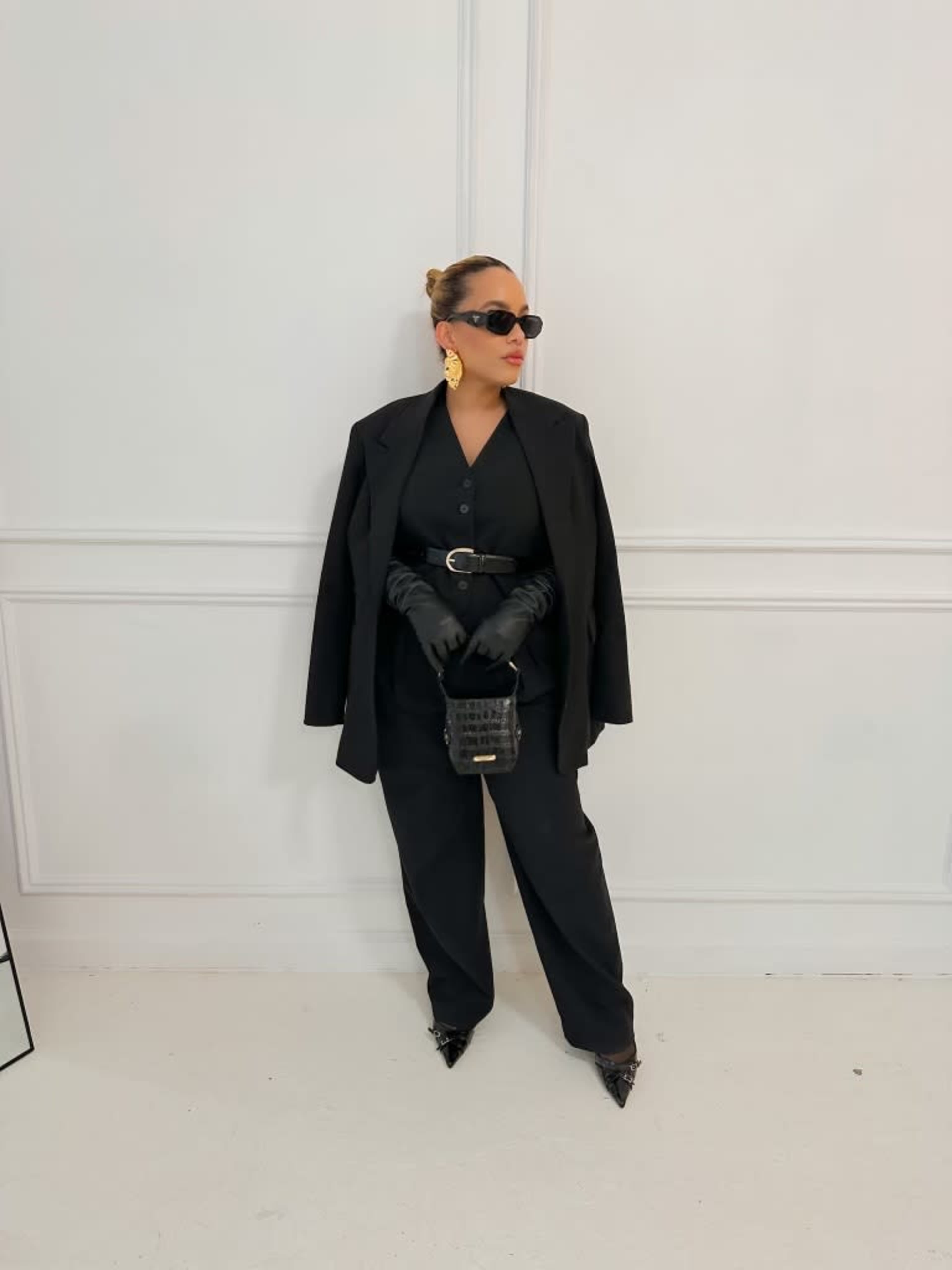 A woman dressed in black clothing, sunglasses, and gloves stands against a plain white wall.