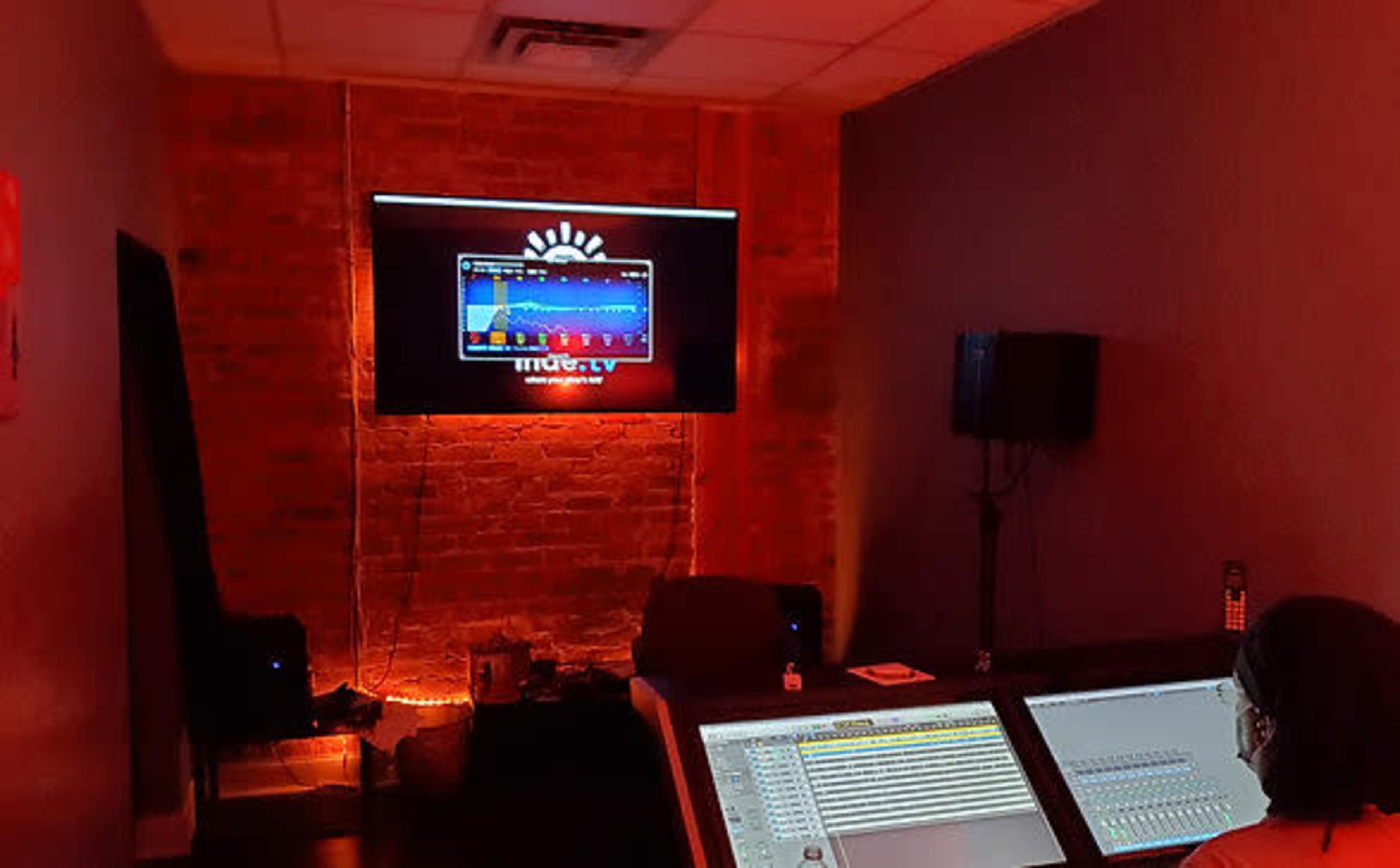 A person is seated in a dimly lit room, focused on a sound mixing console with a large screen displaying audio wave patterns in the background.