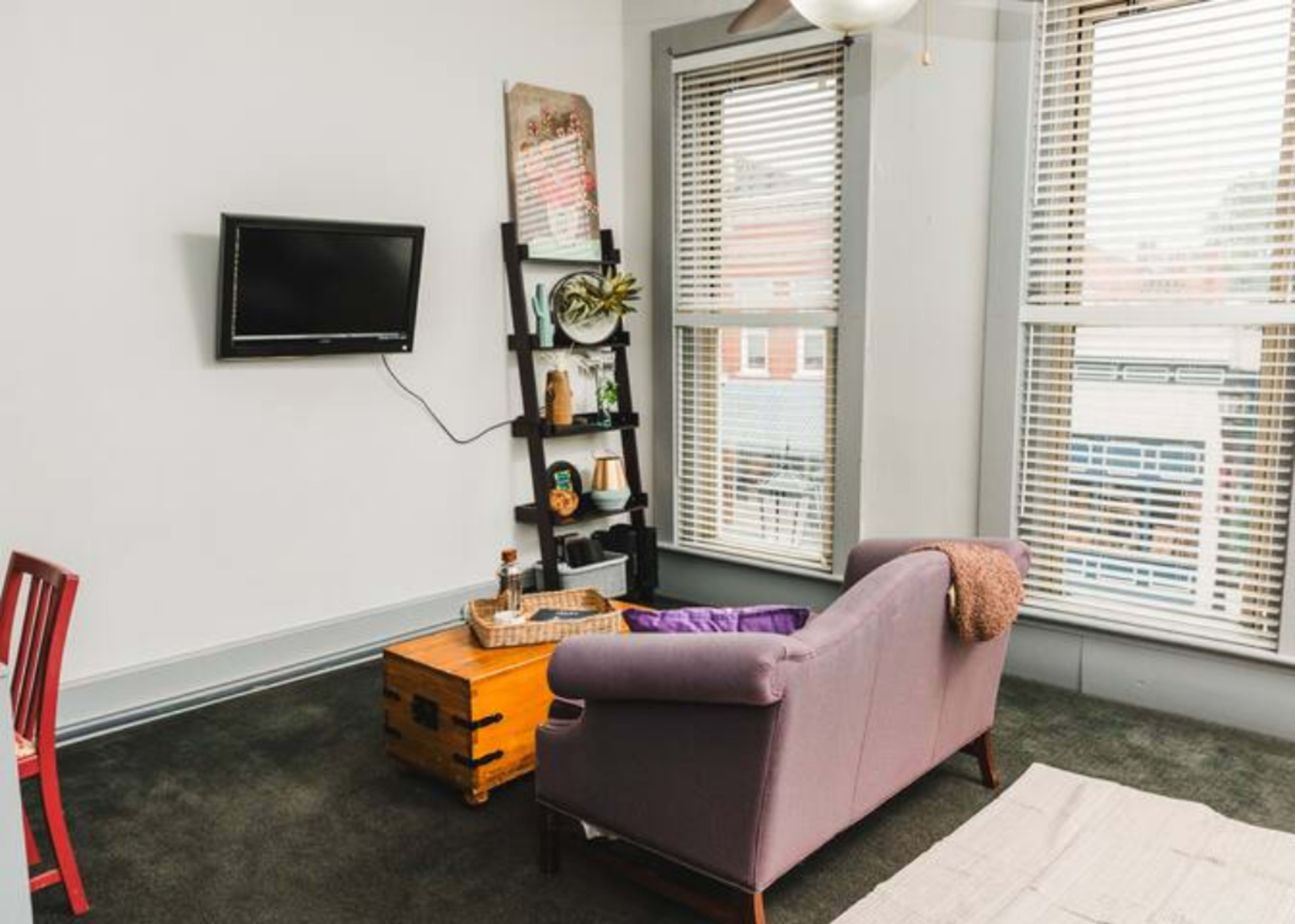 The image shows a living room with a purple couch, a wooden coffee table, a wall-mounted television, and large windows with blinds.