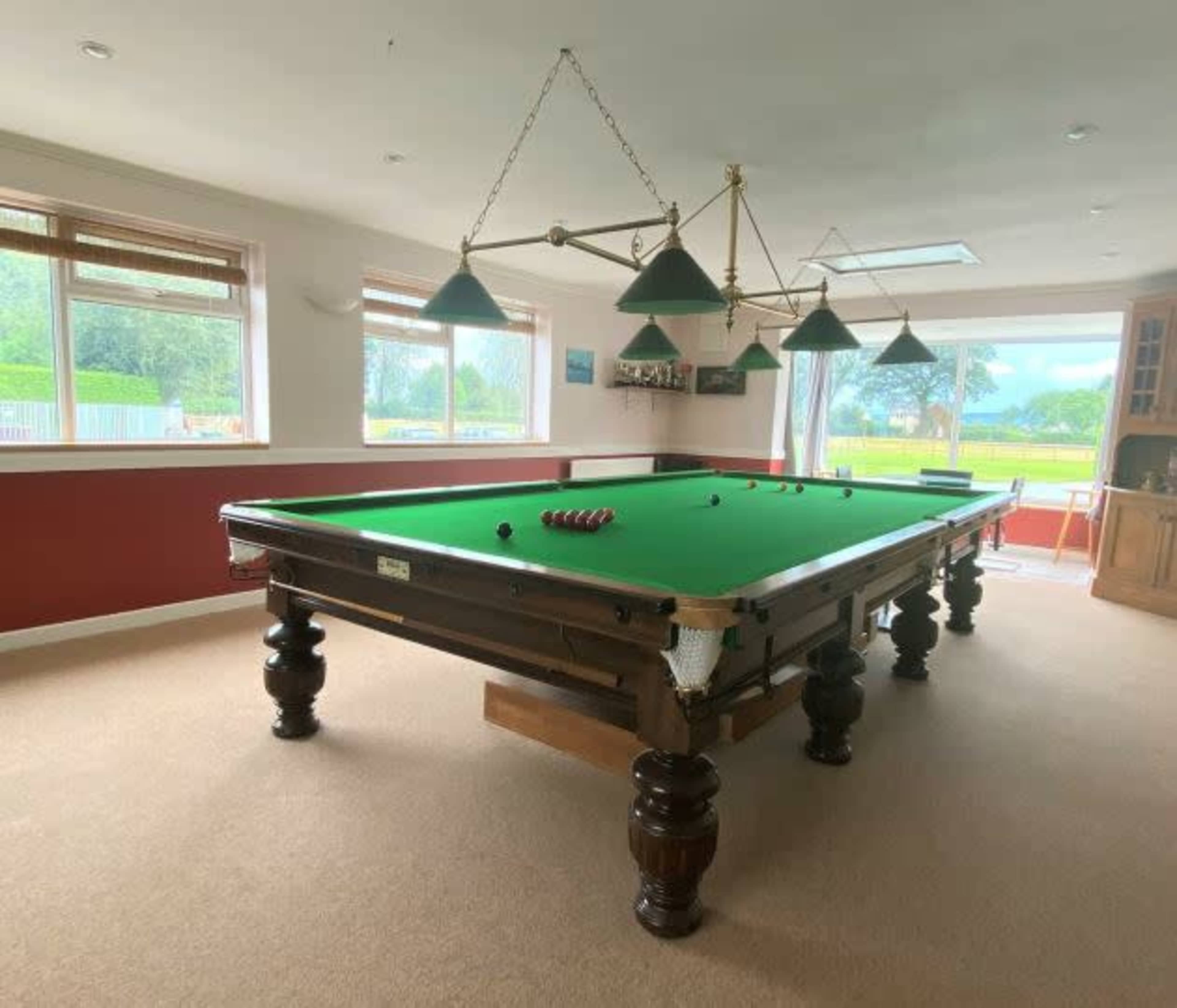The image shows a spacious room featuring a green billiards table surrounded by natural light and decorative elements.