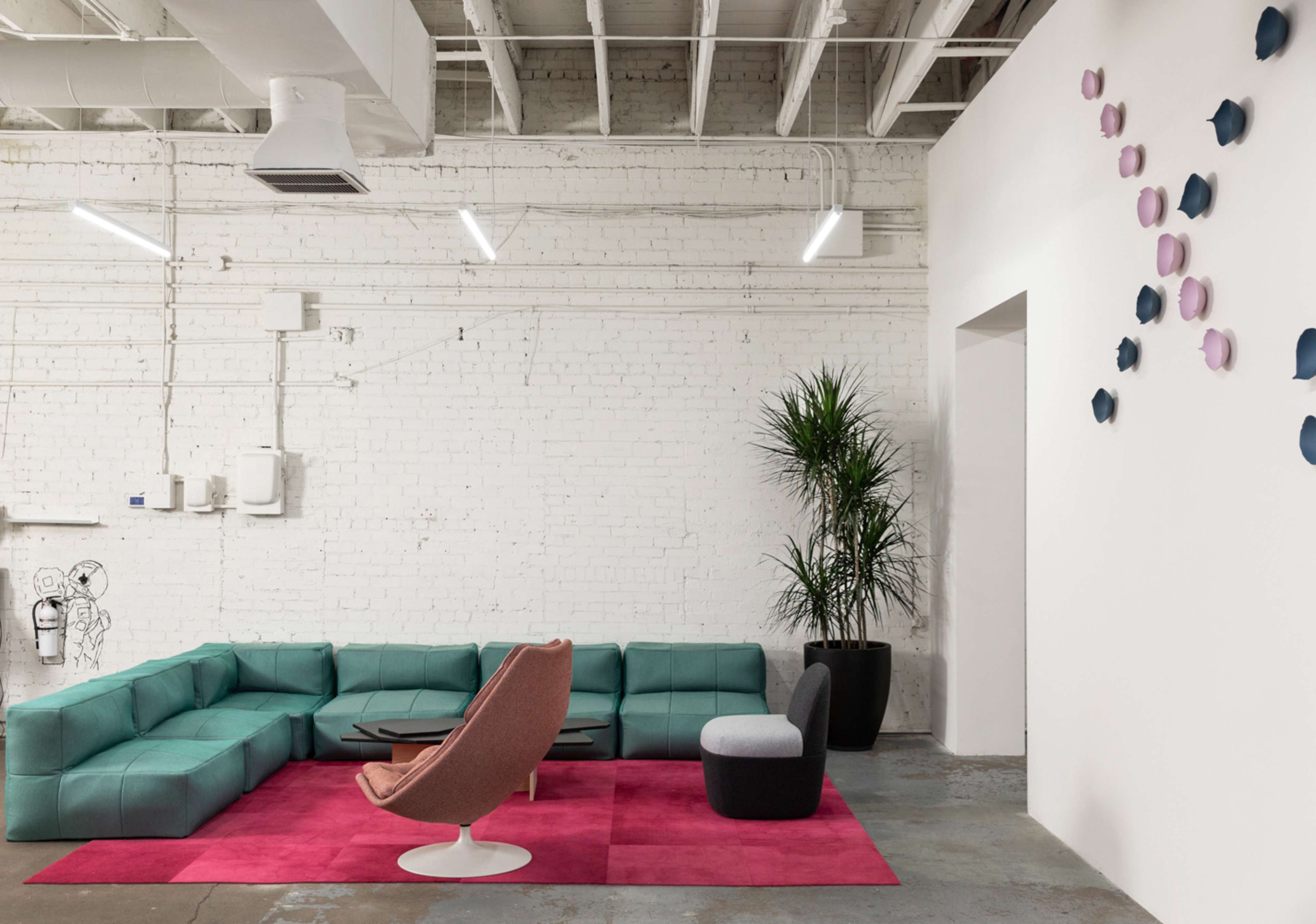 A modern lounge area featuring a turquoise sectional sofa, a pink area rug, a beige chair, and a gray stool, with a potted plant and minimalist wall decor.