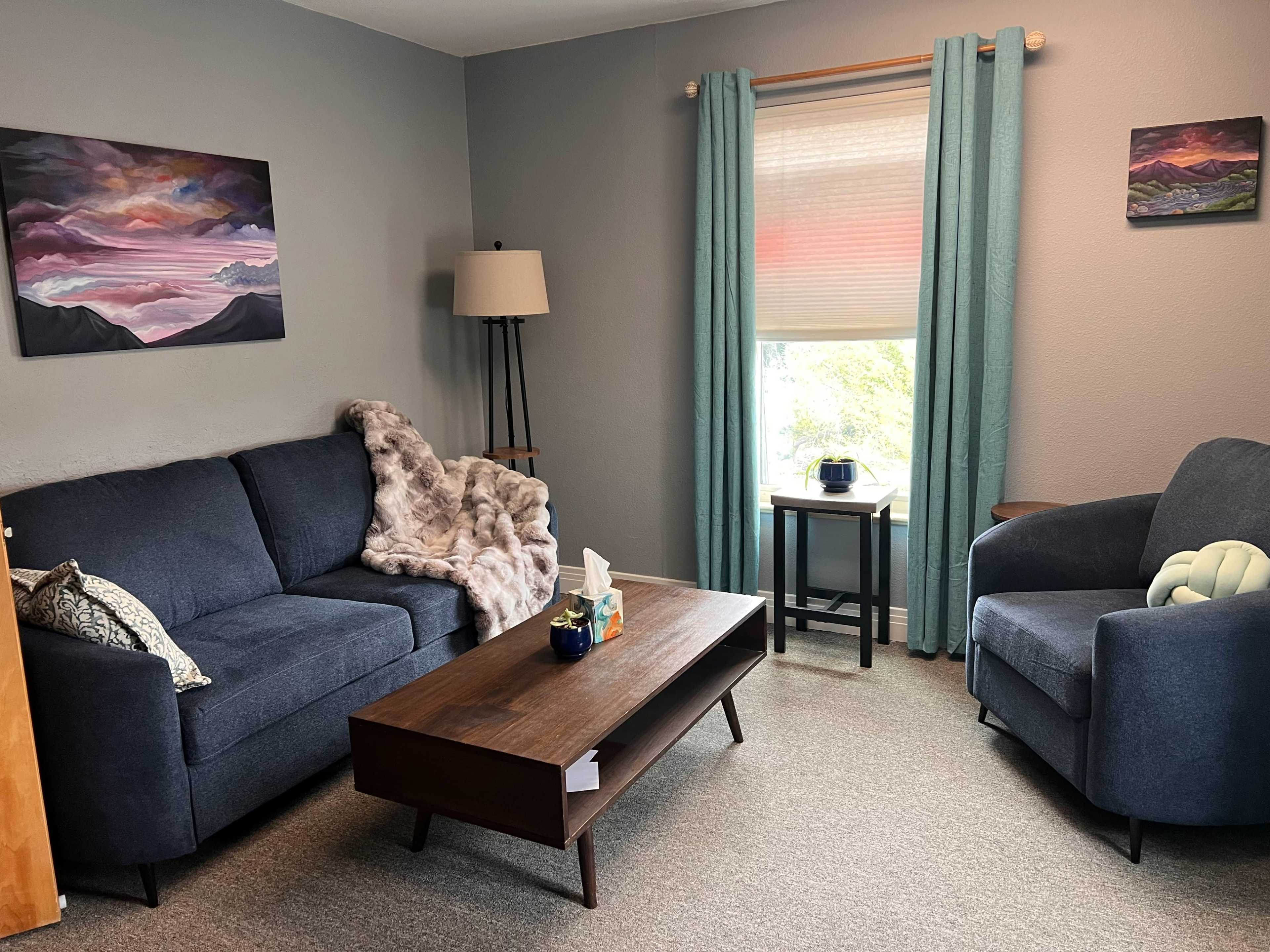The image shows a cozy living room featuring a blue sofa and armchair, a wooden coffee table, and artwork on the walls.