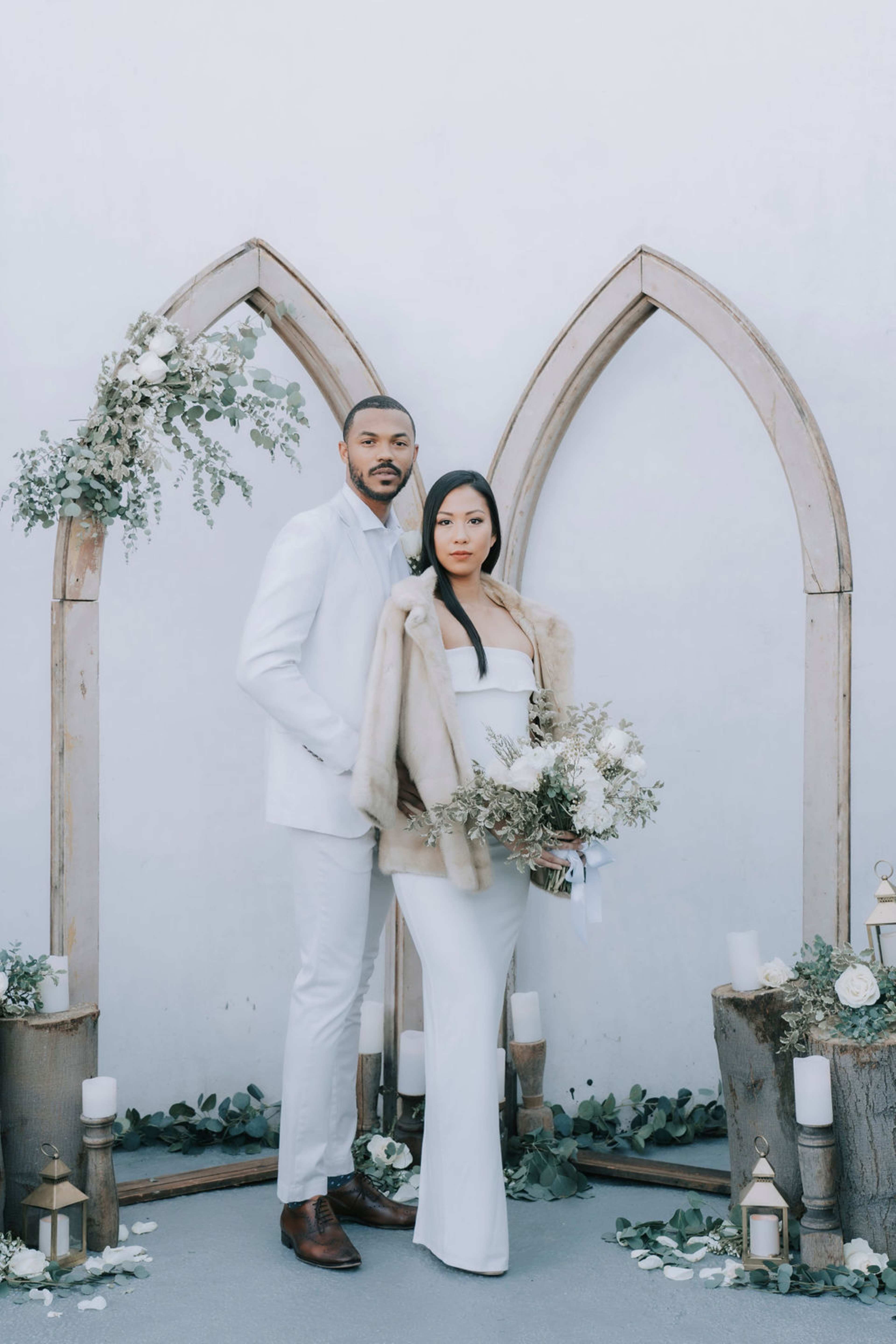 A couple stands in front of a decorative wooden arch adorned with greenery and white candles, surrounded by floral arrangements.