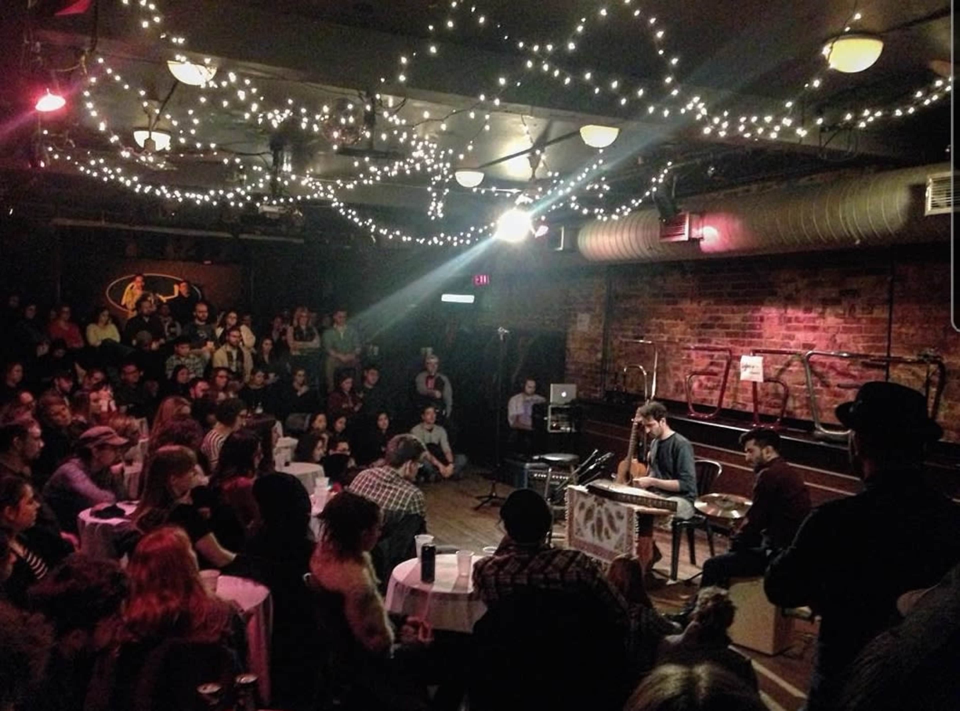 A musician performs on stage in a crowded venue illuminated by string lights.