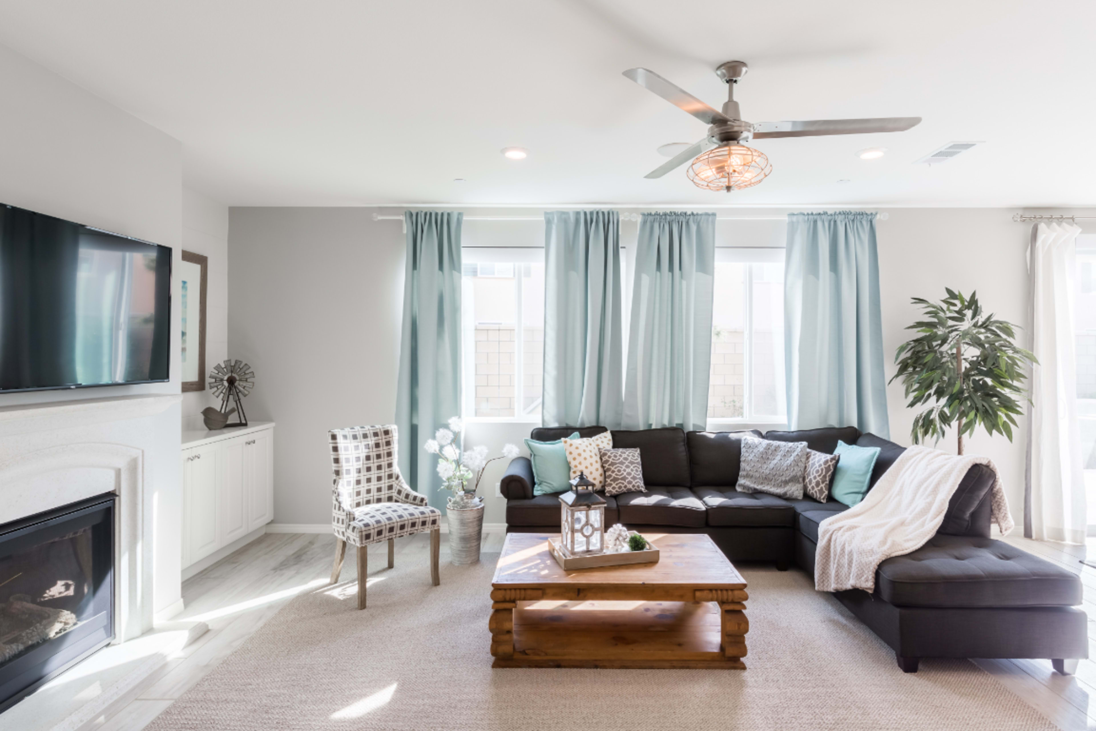 A modern living room features a sectional sofa, a wooden coffee table, a fireplace, and large windows with light blue curtains.