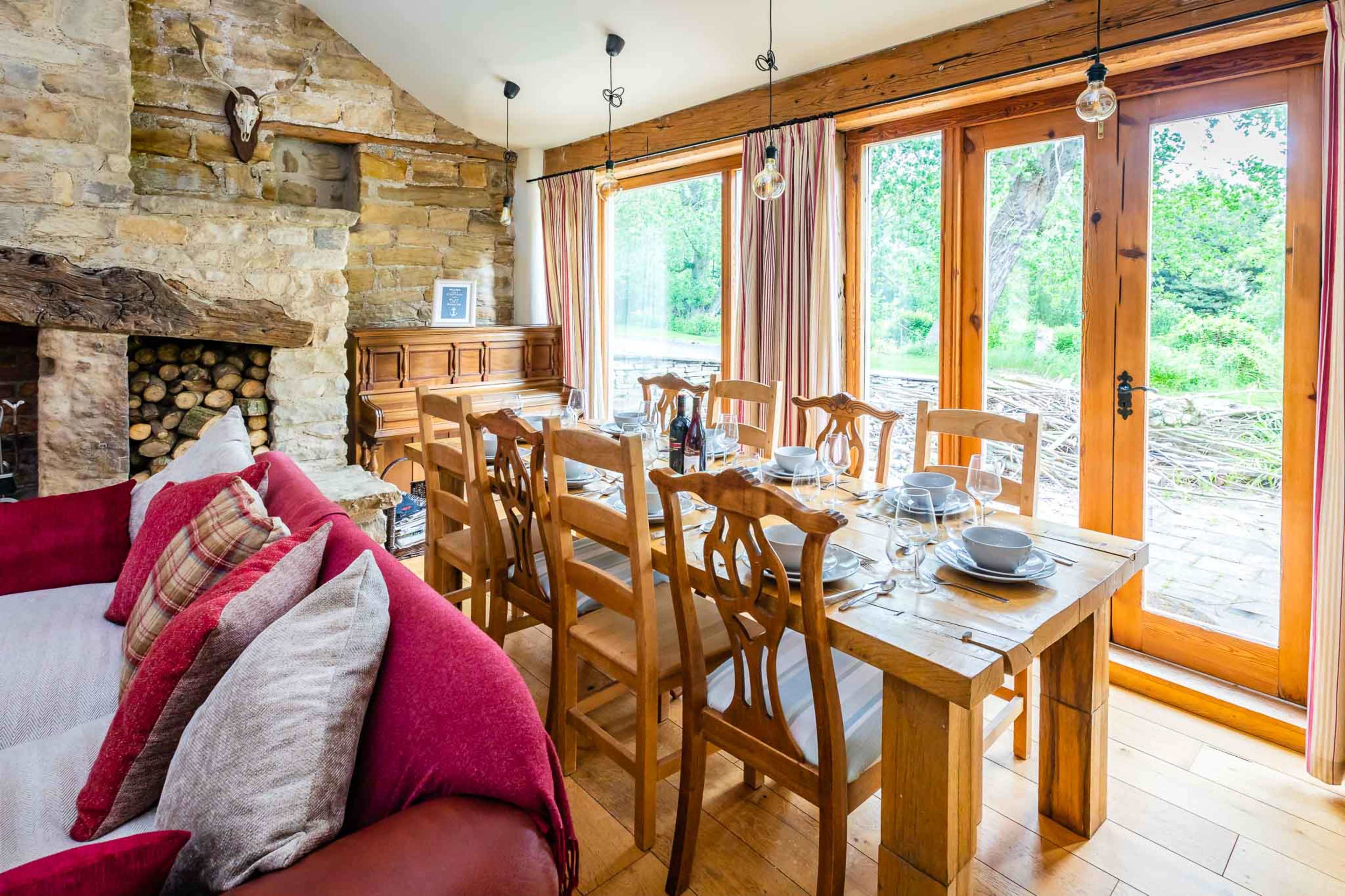 A dining area with a wooden table set for meals, surrounded by chairs, and a cozy seating area with a red sofa overlooking a garden through large windows.