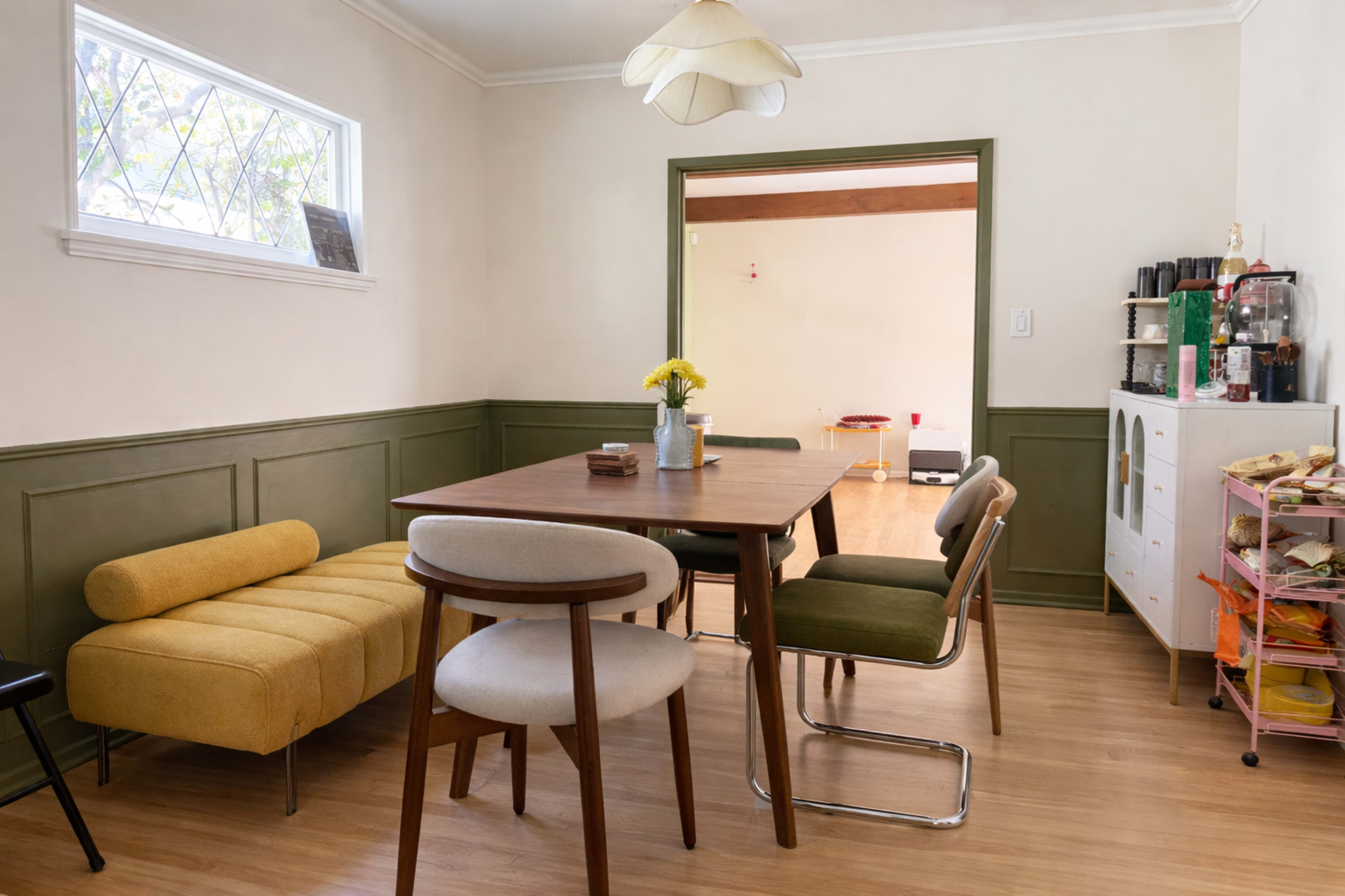 The image shows a simple dining area with a wooden table surrounded by four chairs, a yellow sofa, and a side cabinet, all set against light-colored walls and a green wainscoting.