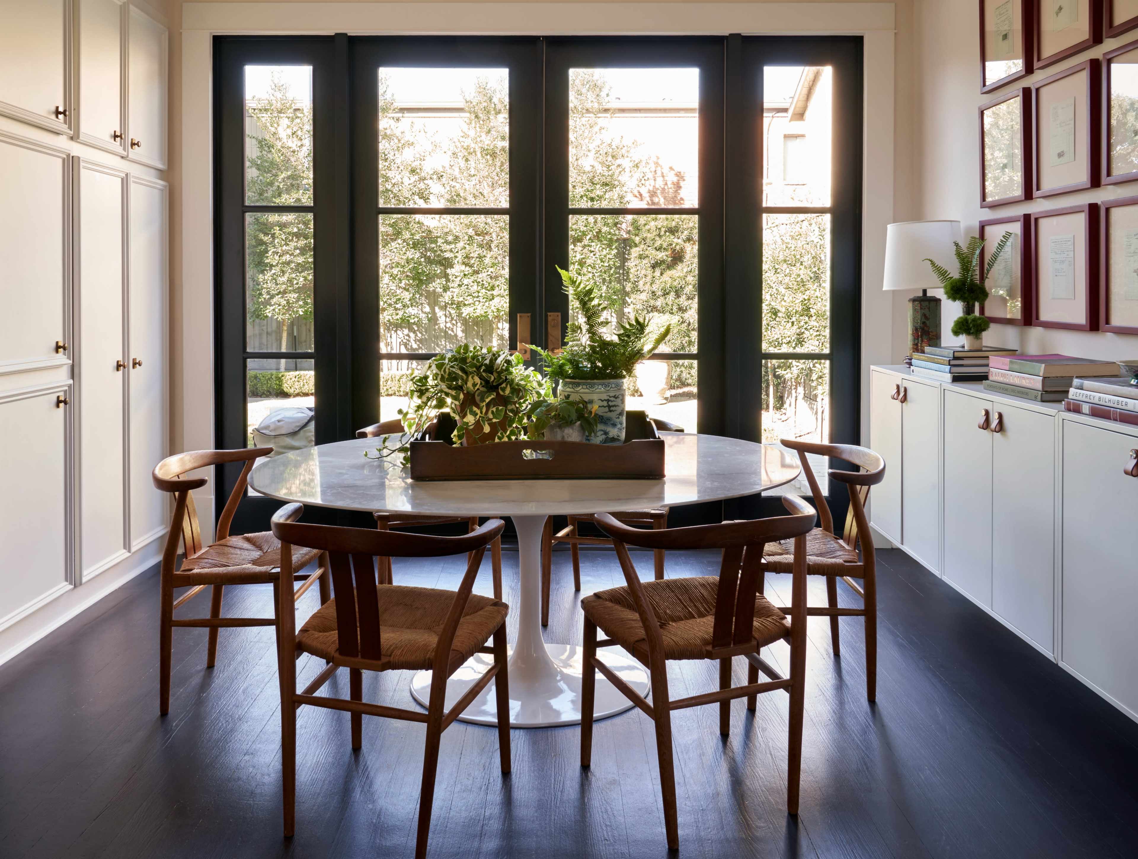 A round dining table surrounded by wooden chairs sits in a well-lit room with large windows overlooking an outdoor garden.