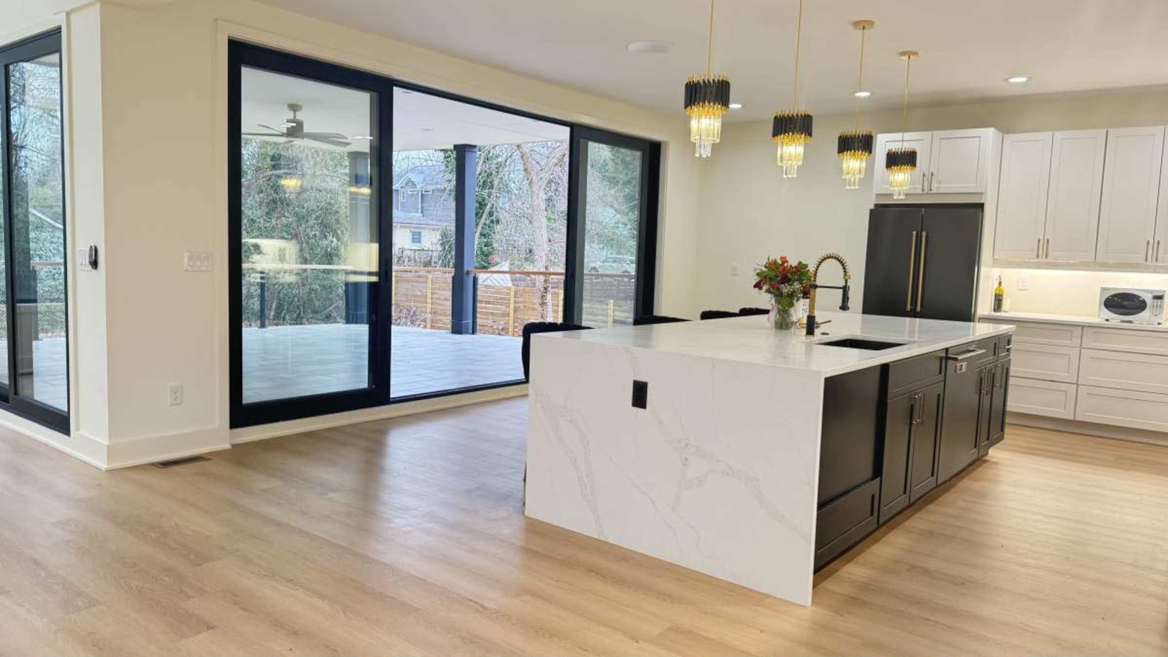 The image shows a modern kitchen with a large island, black cabinetry, and a connected dining area featuring large glass doors that lead to a deck.